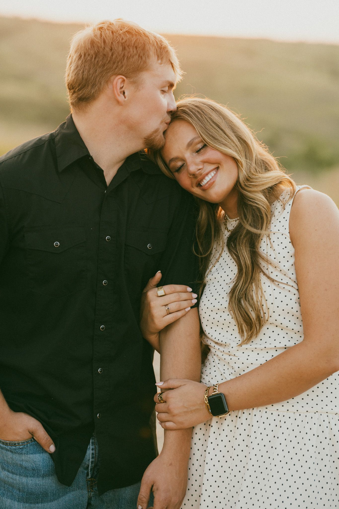 A couple shares a tender moment outdoors, man in black shirt kissing womans cheek as she smiles in white polka dot dress.