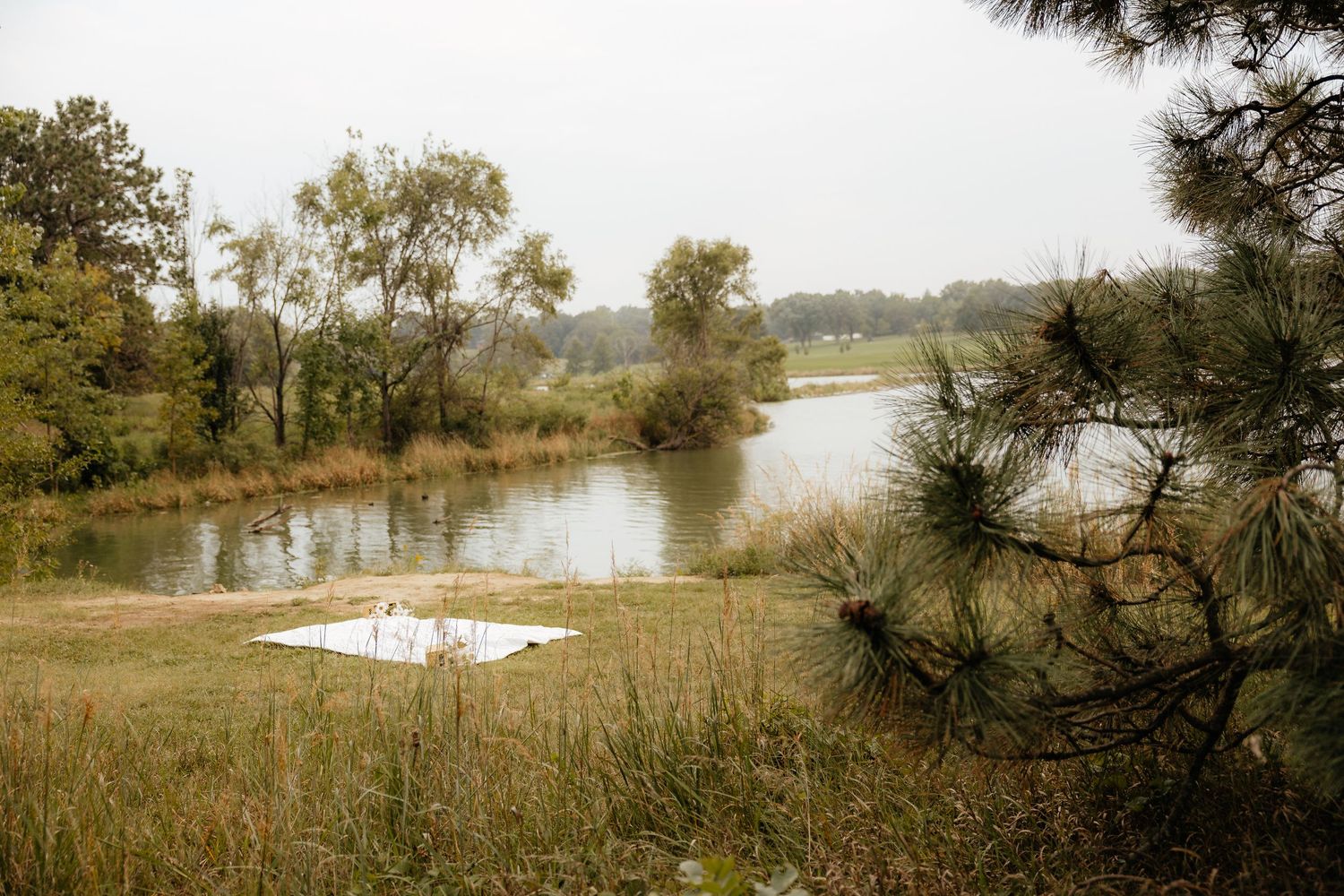 A white picnic blanket spread on grass beside a calm river, surrounded by autumn trees on a cloudy day.