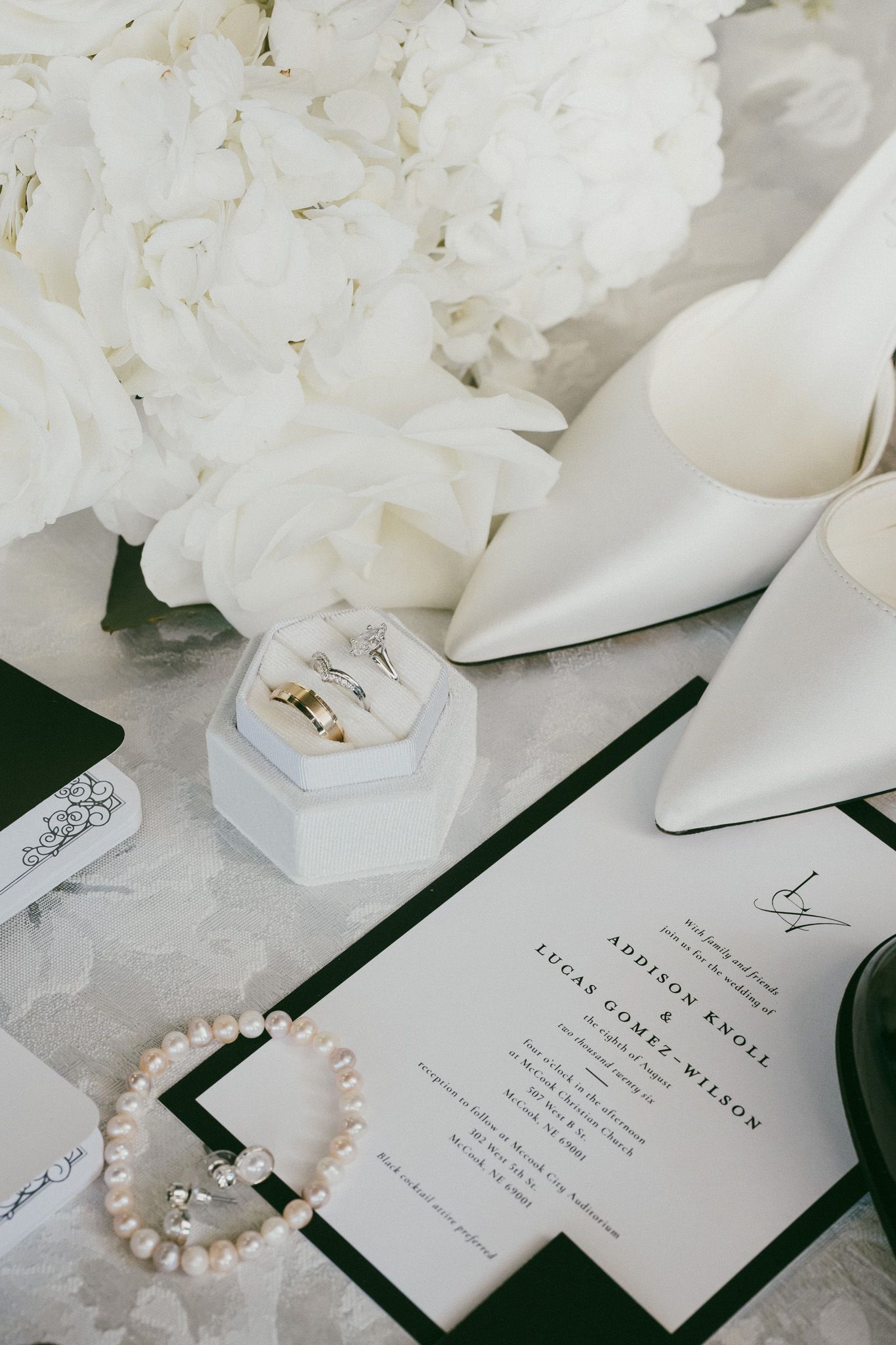 Elegant wedding flat lay with white hydrangeas, pearl bracelet, gold rings, ivory heels, and green-trimmed invitation suite.