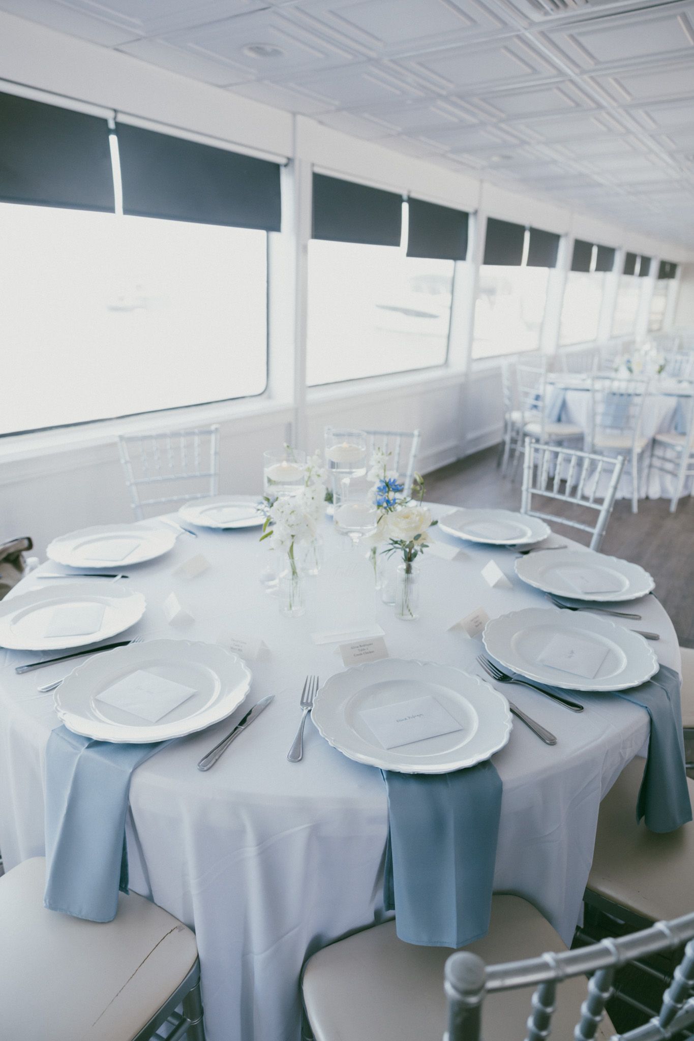 Elegant wedding reception table set with light blue linens, white plates, and floral centerpieces in a bright venue.