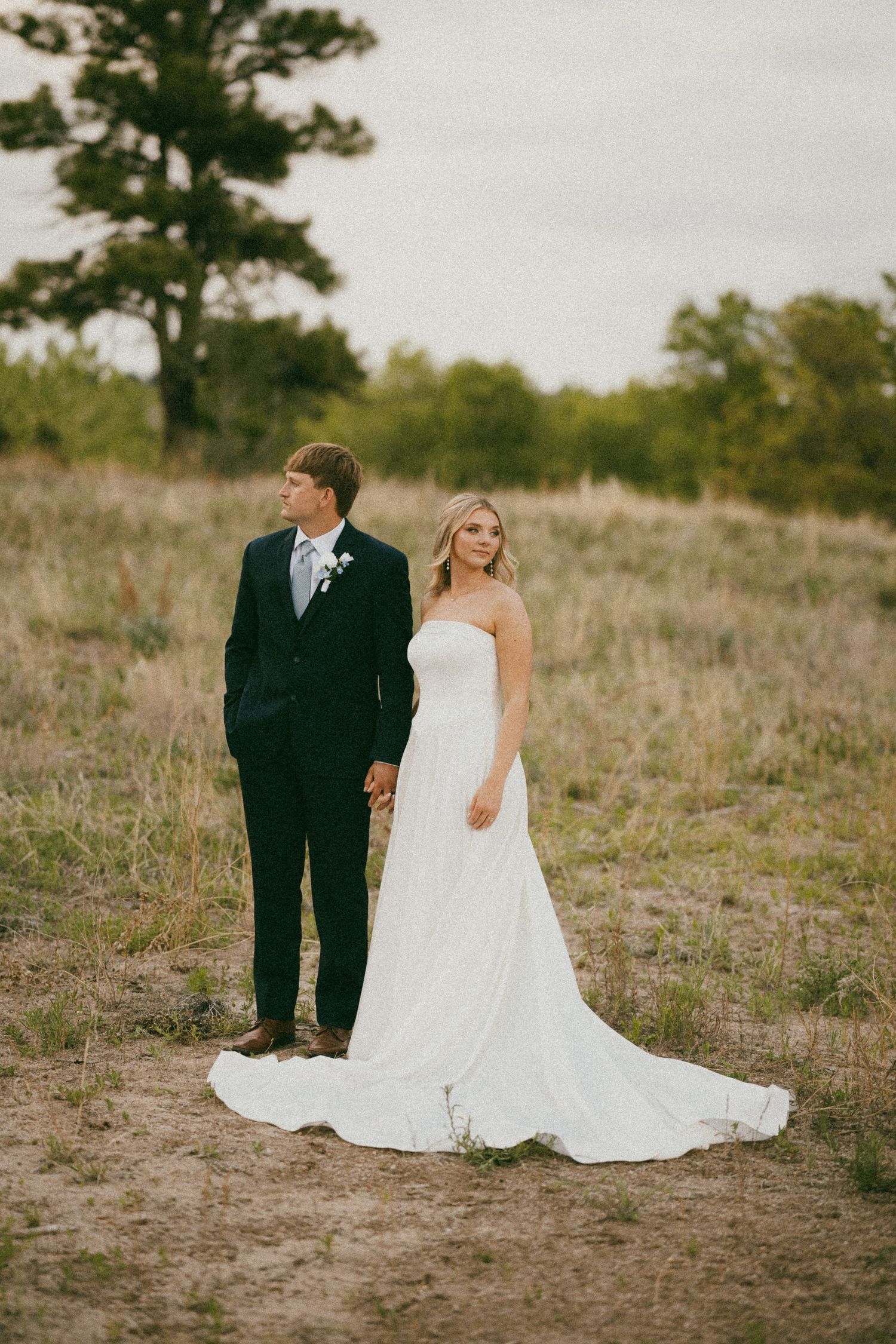 Bride in strapless white gown and groom in dark suit hold hands in rustic outdoor field during wedding portrait.