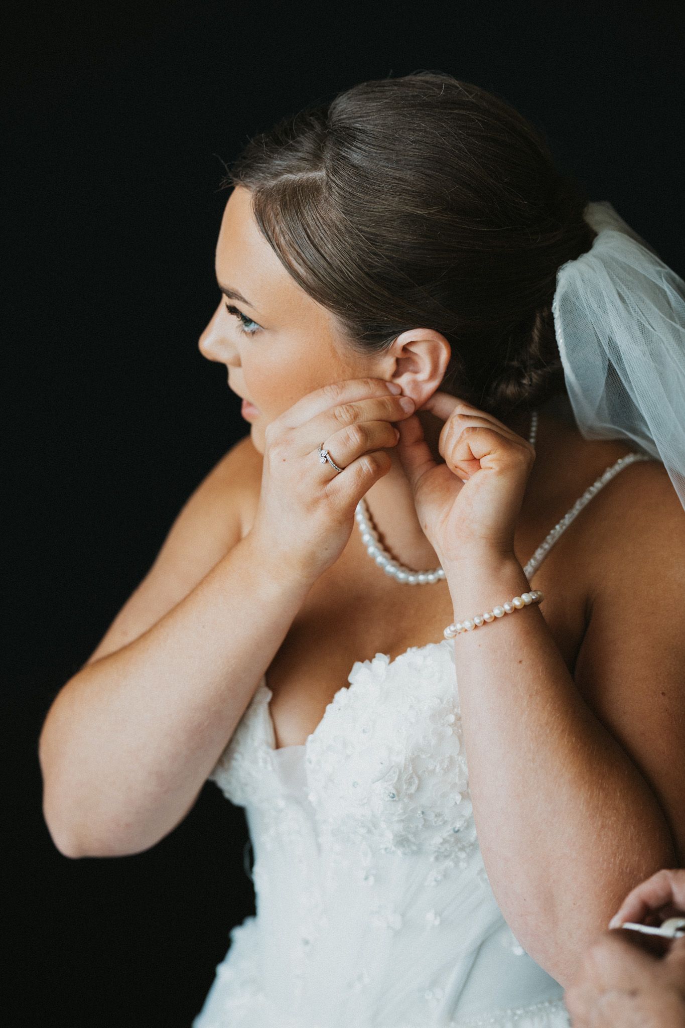 Bride in white strapless gown puts on pearl earring, wearing veil and pearl necklace, against dark background.