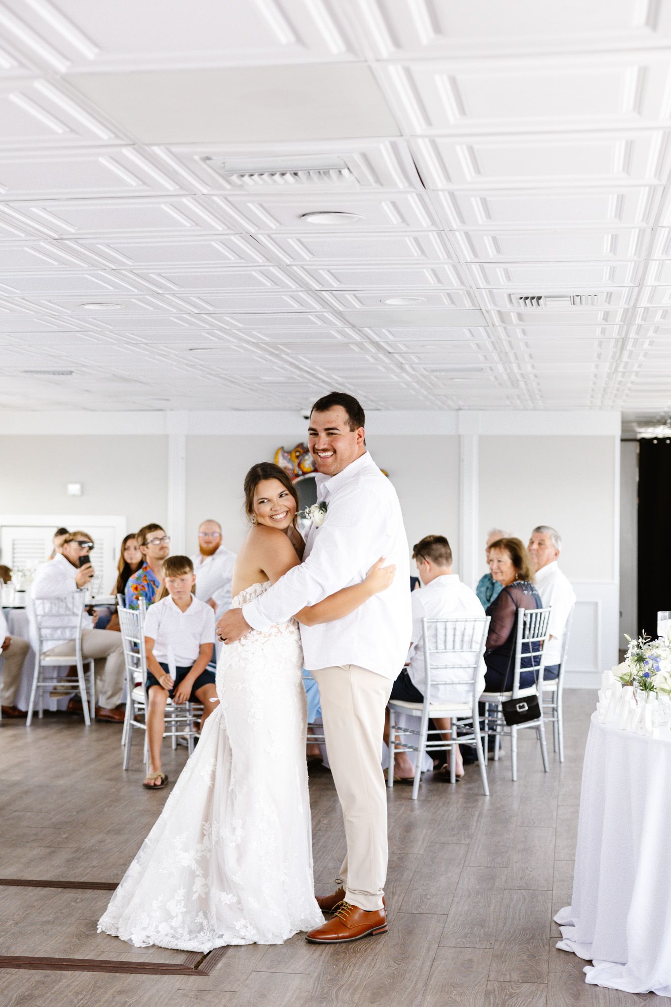 Bride and groom share first dance at elegant white wedding reception venue with guests seated at tables.
