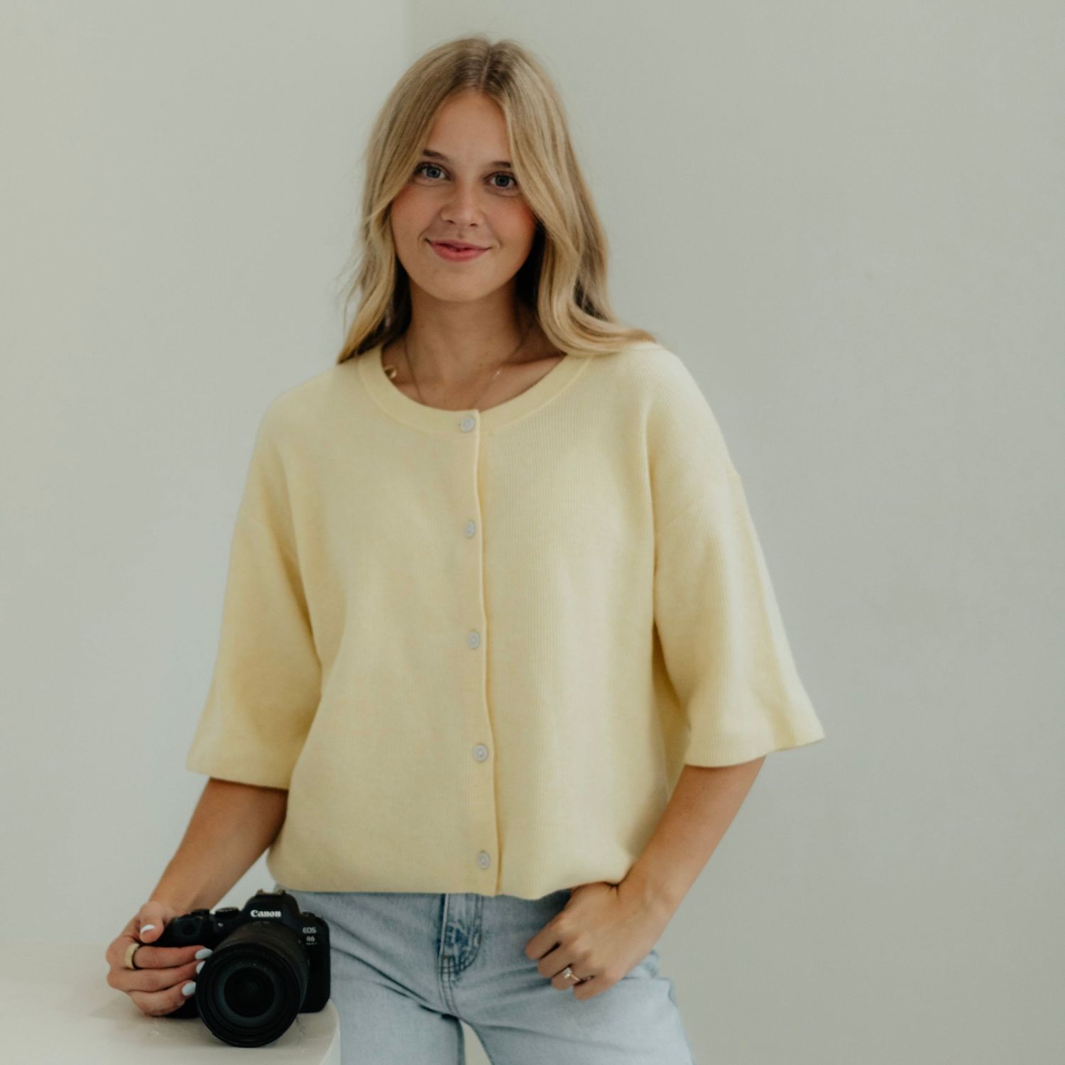 Woman in cream cardigan holding a Canon camera, smiling against a light gray background.