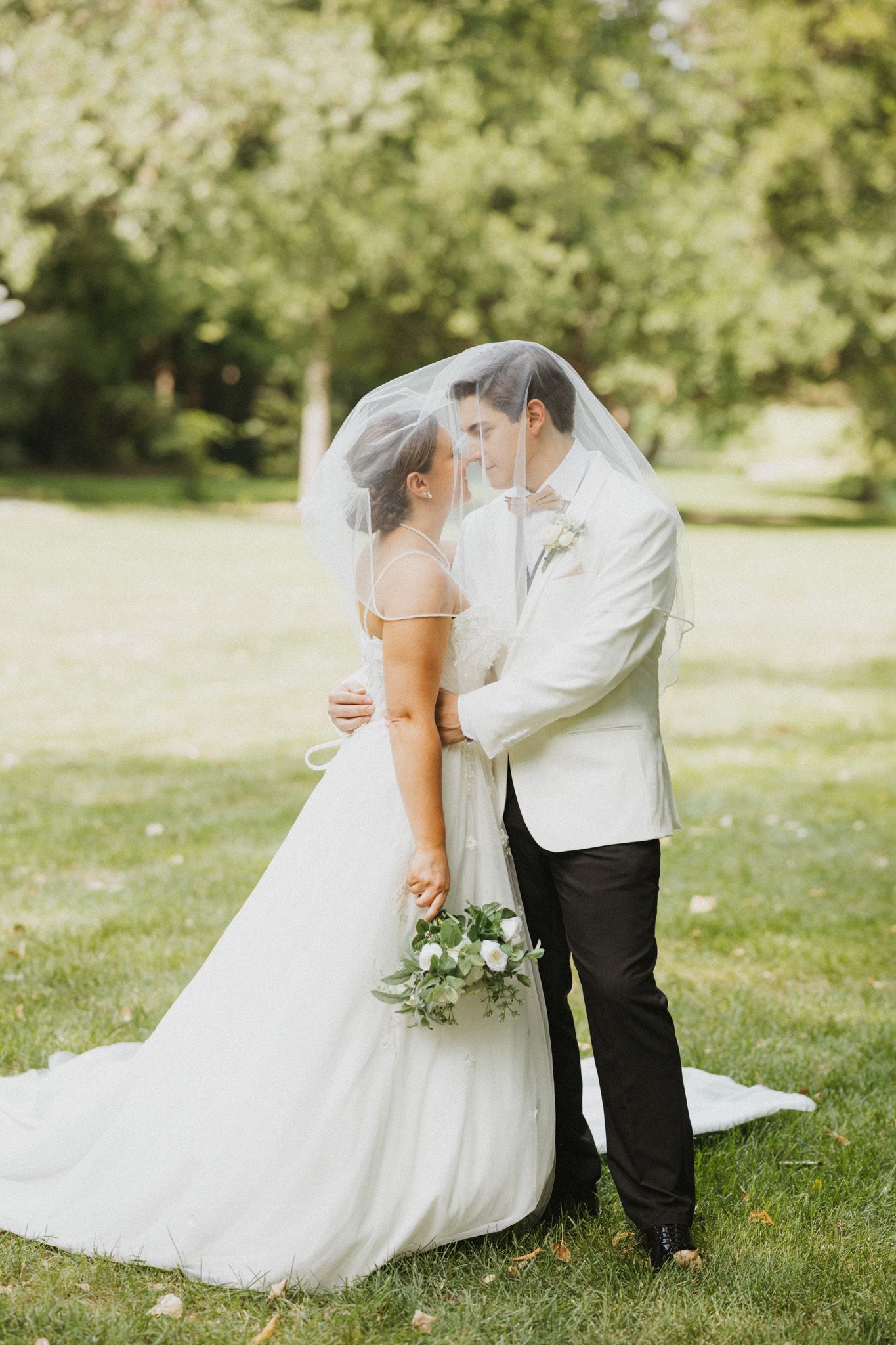 Bride and groom share a romantic moment under a veil outdoors, bride holding white bouquet in lush green park.