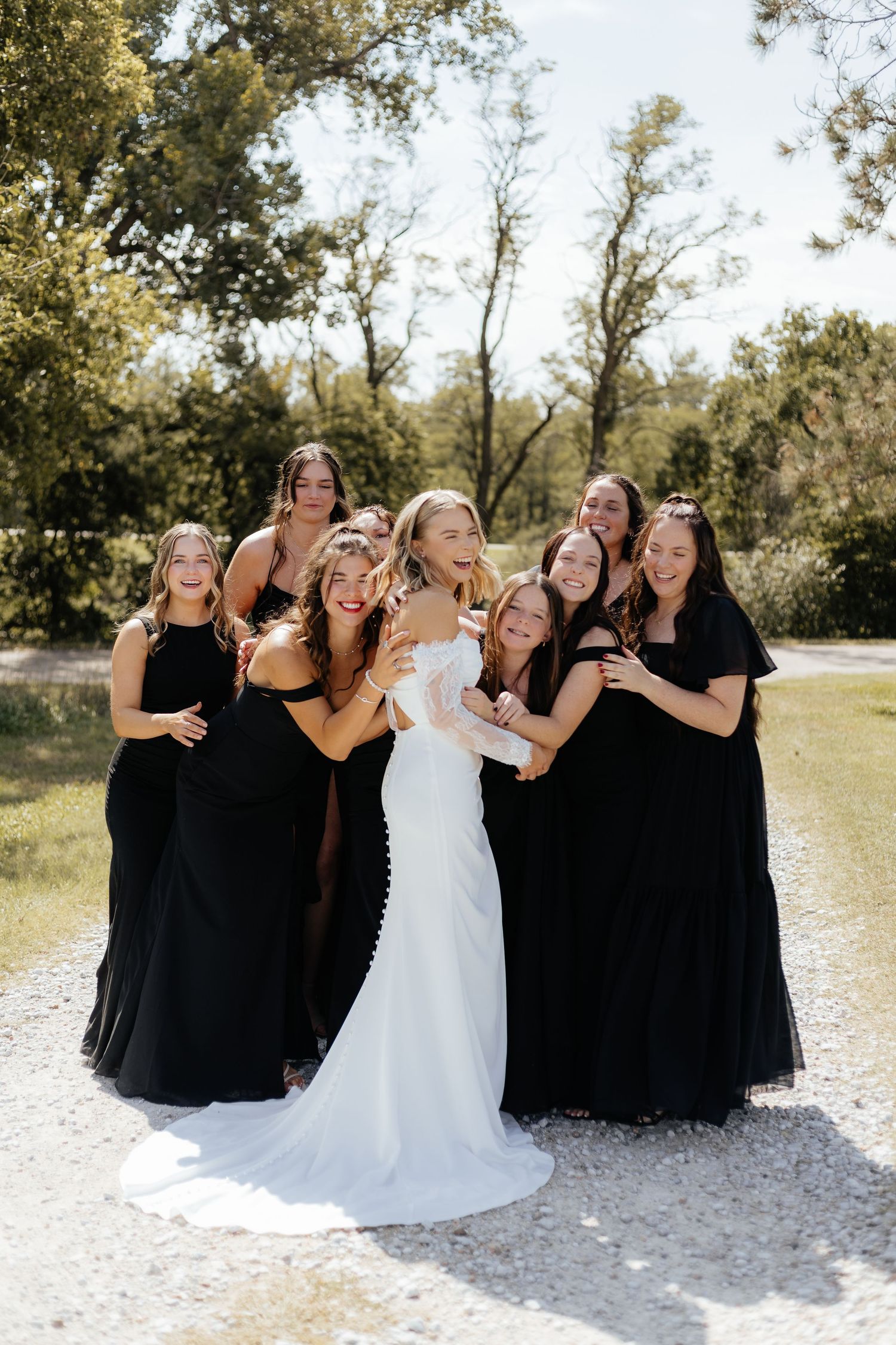 Bride in white gown surrounded by bridesmaids in black dresses, laughing outdoors on a sunny day.