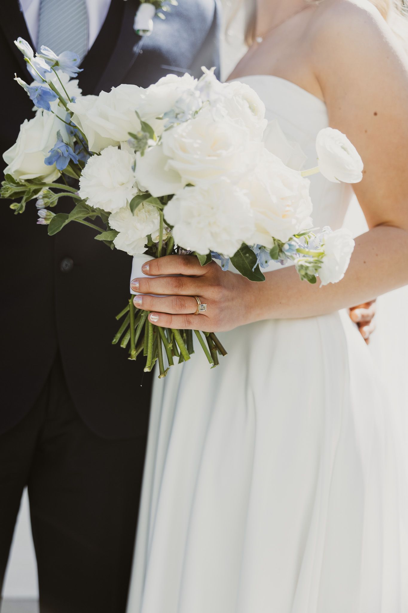 Bride holding white and blue wedding bouquet of roses and delphinium while standing next to groom in navy suit.