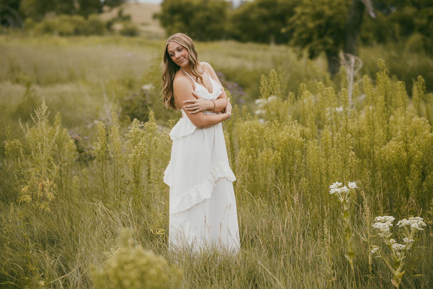A woman in a white flowing dress stands gracefully in a lush green meadow field with wildflowers.