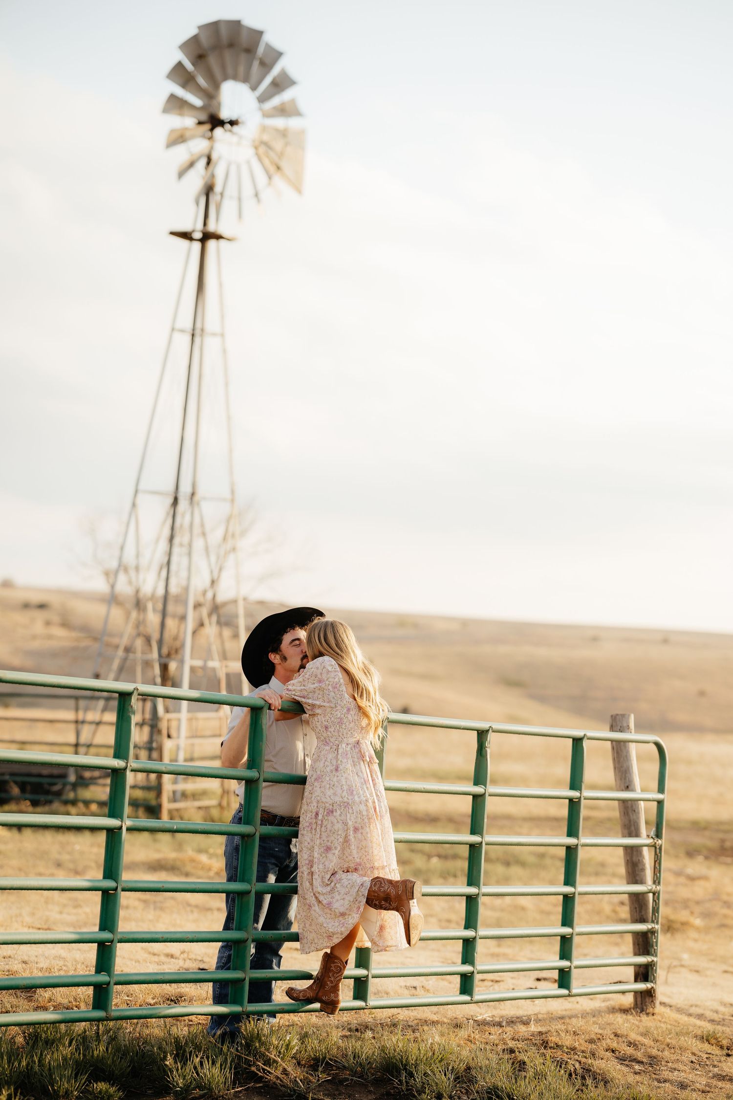 Couple kissing by a green farm gate with a windmill in the background on a sunny rural landscape.