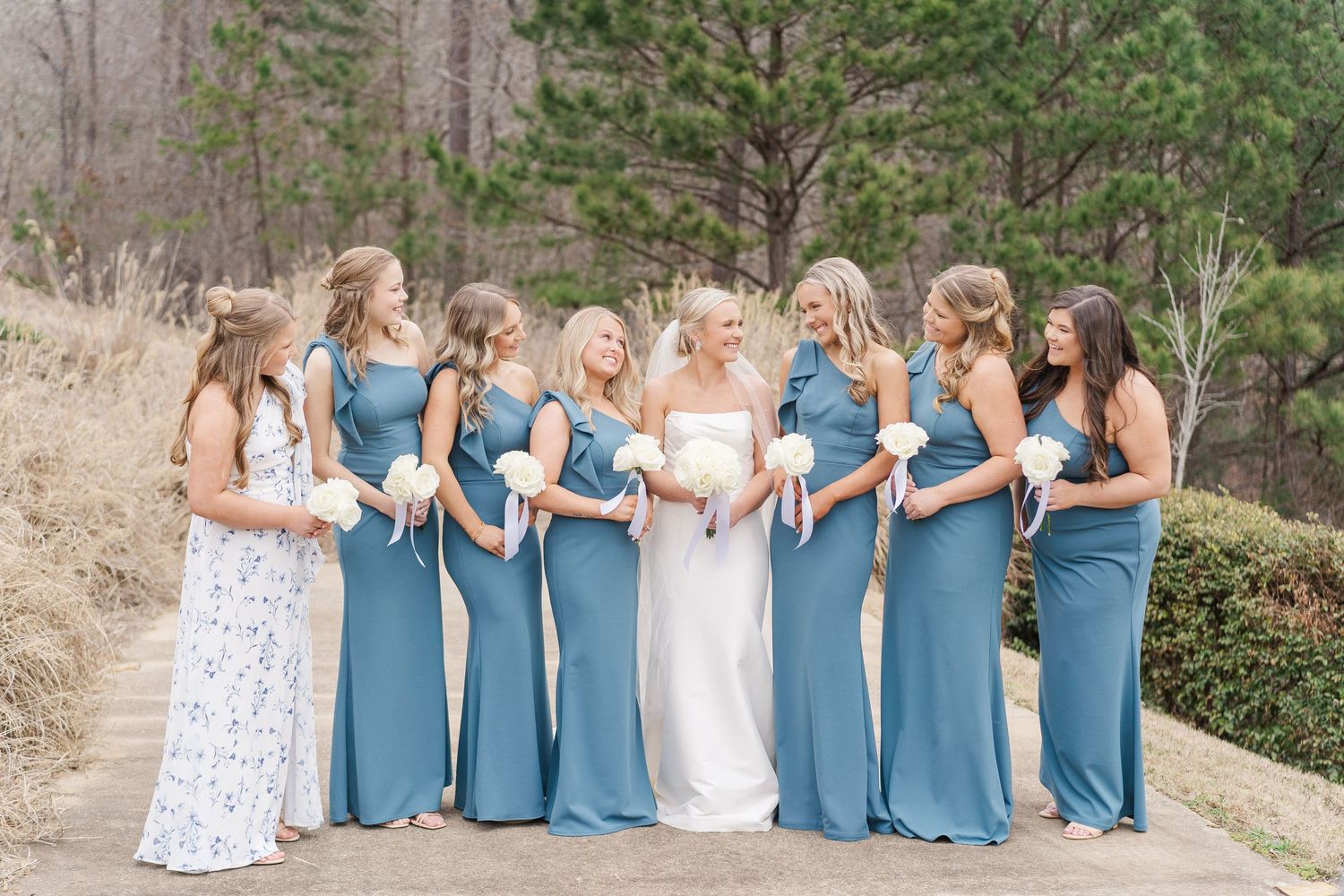 Wedding party wearing coordinated dusty blue bridesmaid dresses stands together on a hillside path in natural setting.