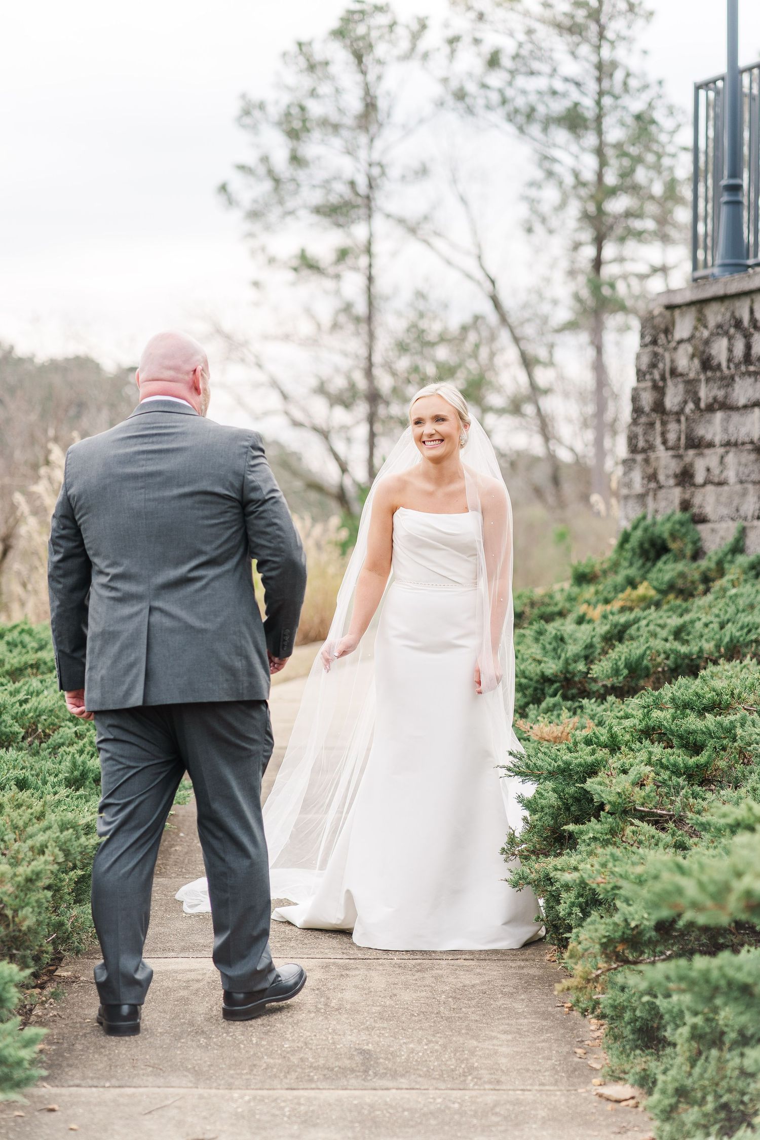 A bride in a flowing white strapless gown stands on a garden path as her groom faces her during their first look moment.