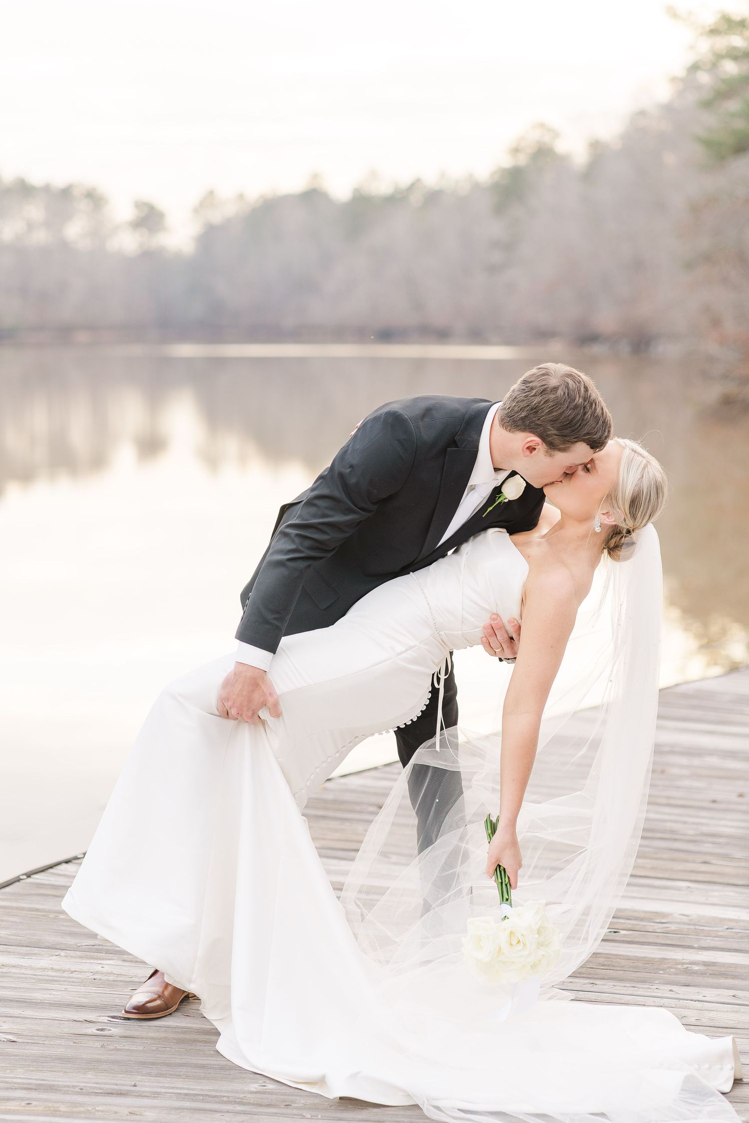 Romantic wedding couple dips for a kiss on wooden dock by misty lake at sunset with flowing white veil.