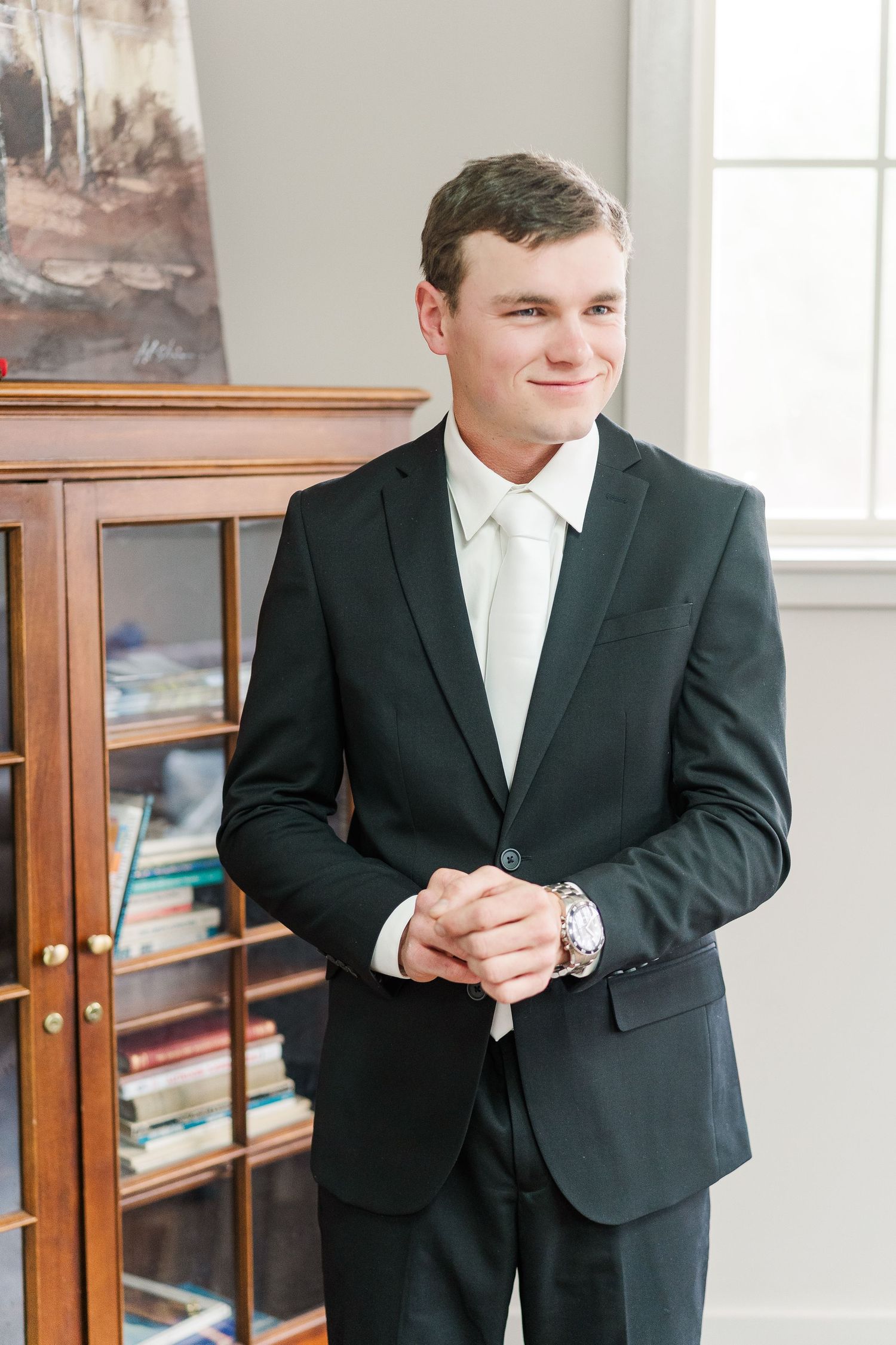 A person in a classic black suit and white tie stands poised beside a wooden display cabinet in a bright interior setting.