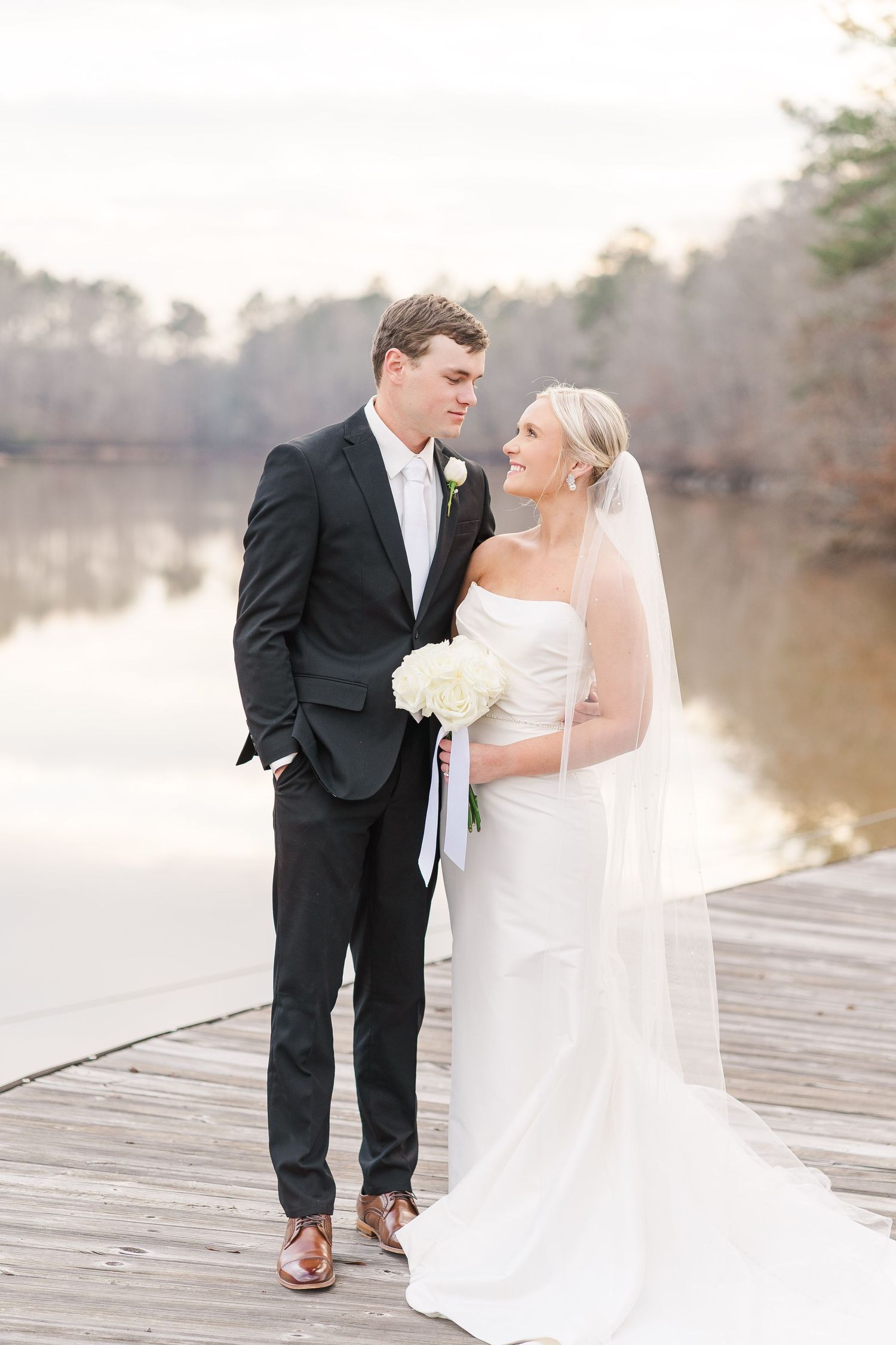 A wedding couple shares an intimate moment on a wooden dock by a serene lake during golden hour.