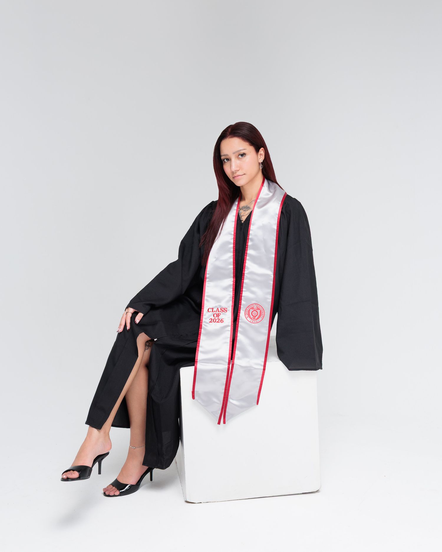 Woman in black graduation gown and white sash sits on white cube, posing for professional grad photo.