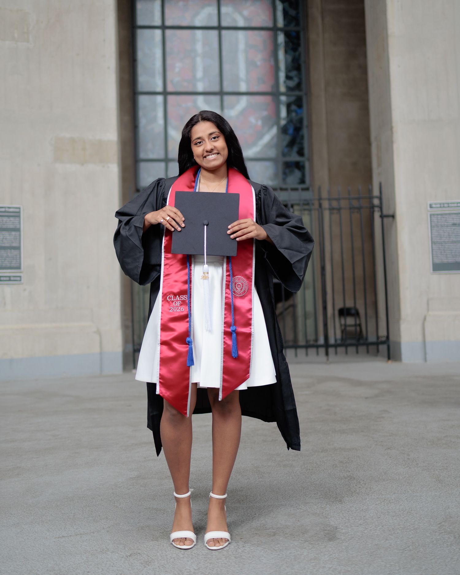 A smiling graduate in black cap and gown with red sash holds diploma outside university building.