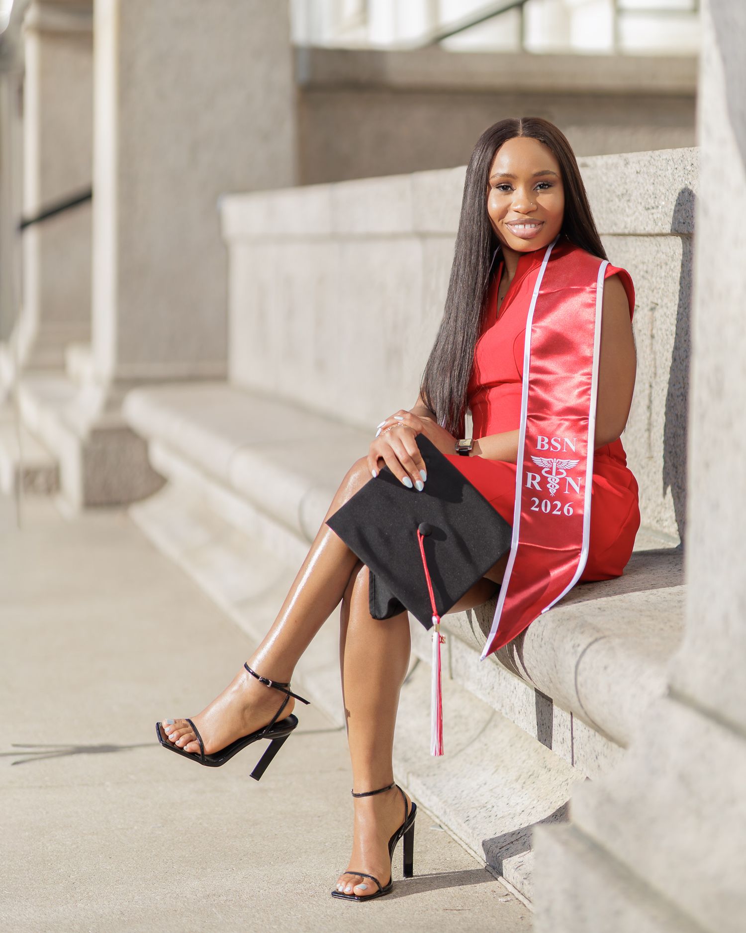A smiling graduate in a red dress poses on stone steps holding a black cap, wearing a BSN RN 2026 sash.