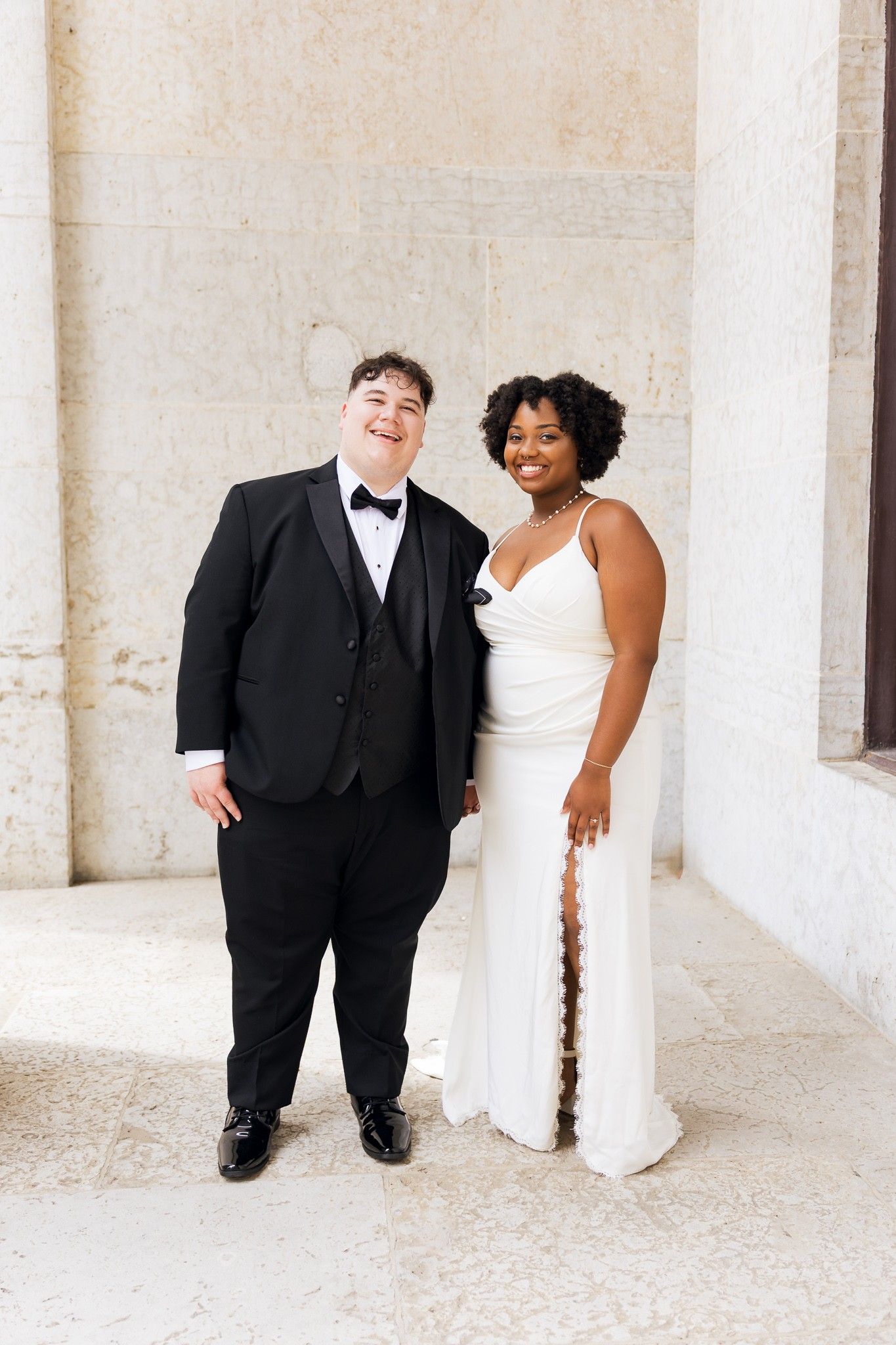 Couple in wedding attire, one in black tuxedo and one in white gown, smiling together outdoors.