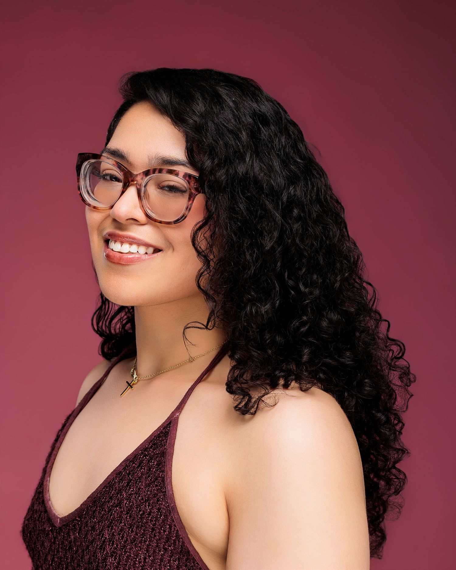 Smiling young woman with curly black hair and tortoiseshell glasses wearing a burgundy top against a pink background.