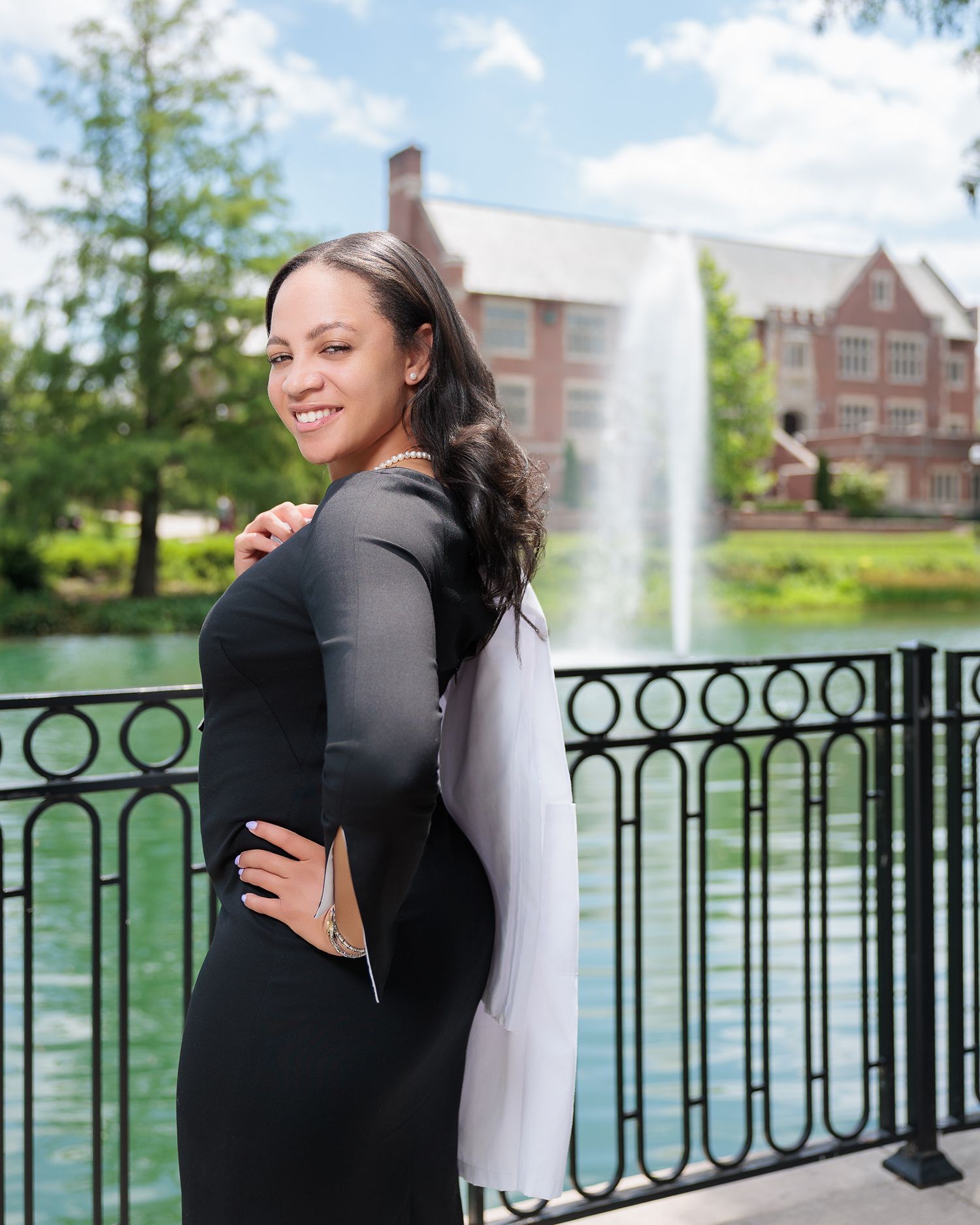 A smiling woman in a black dress poses by a decorative fence with a fountain and brick building in the background.