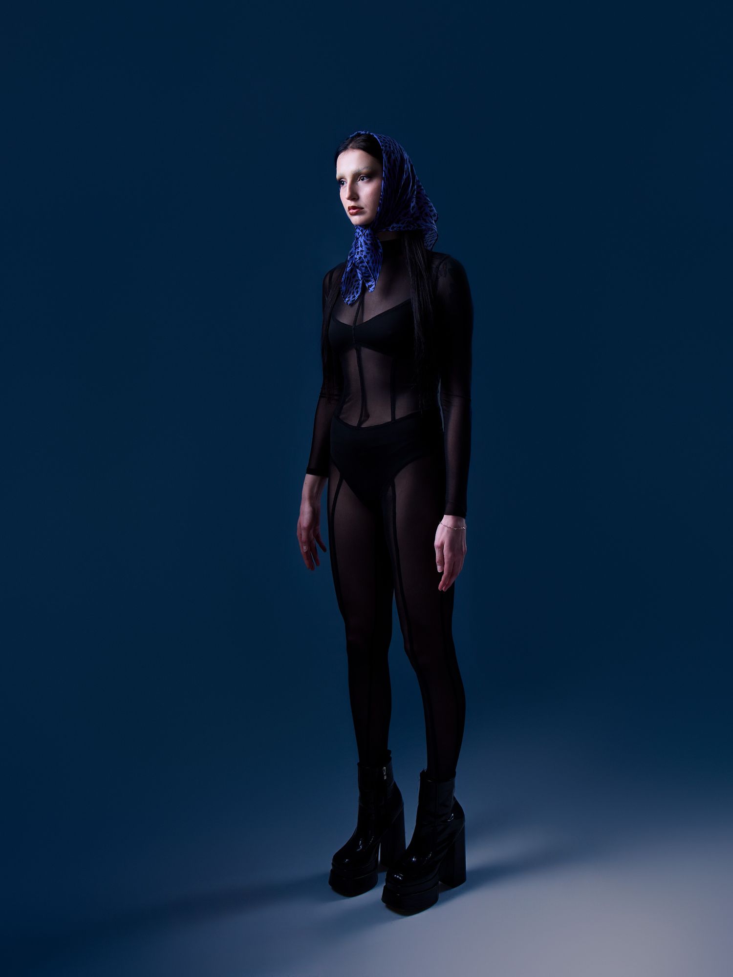Woman in black sheer bodysuit and platform boots with purple hair against dark blue background.