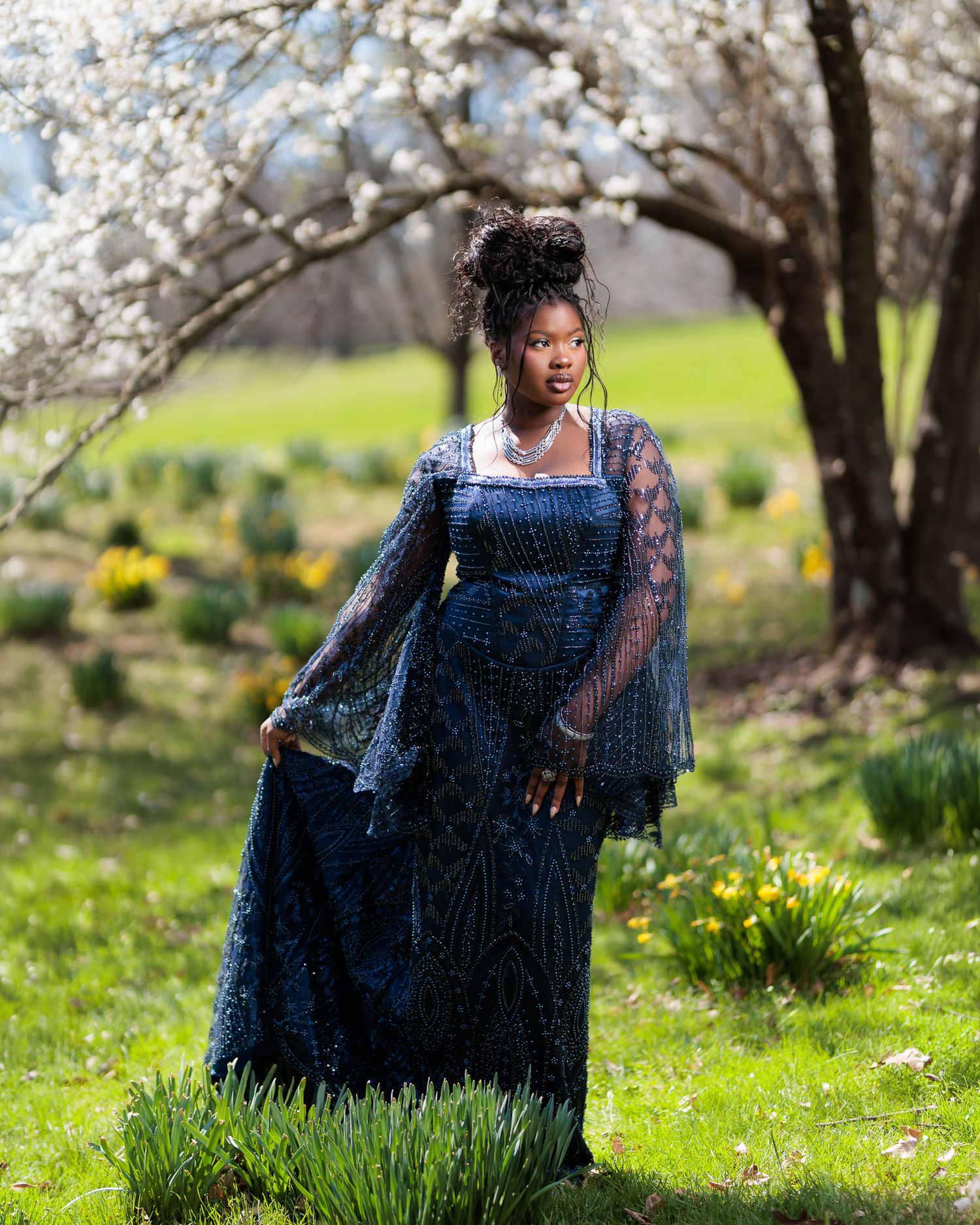 Woman in navy beaded gown poses under blooming white tree surrounded by daffodils in spring garden.