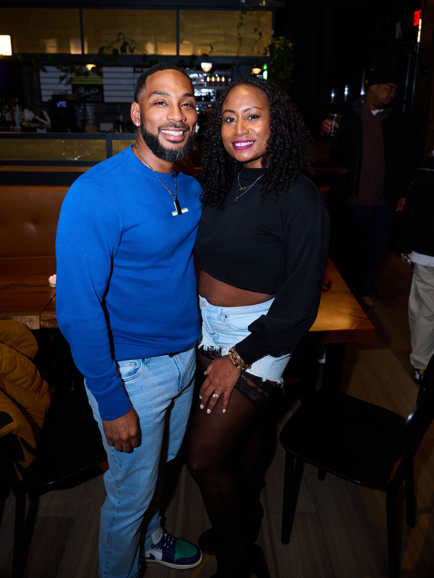 Two people wearing blue and black outfits pose together at a dimly lit venue during an evening event.
