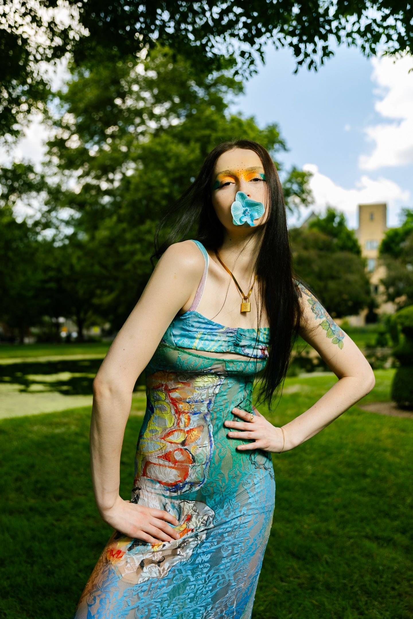 Model in blue multicolor dress blowing blue bubble gum in sunny park with hand on hip.
