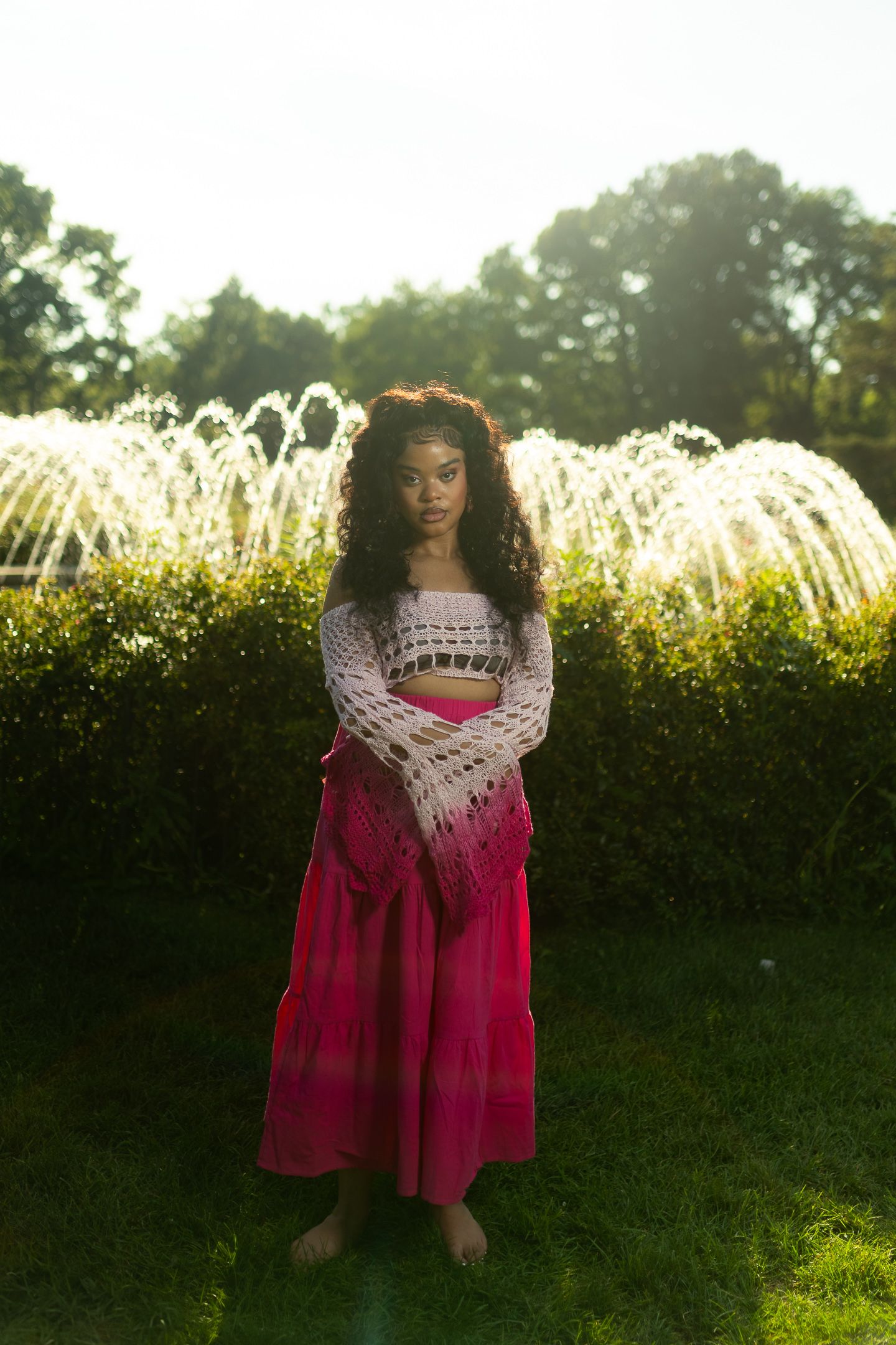 Young woman in pink skirt and crochet top standing barefoot on grass near fountain in sunlit park.
