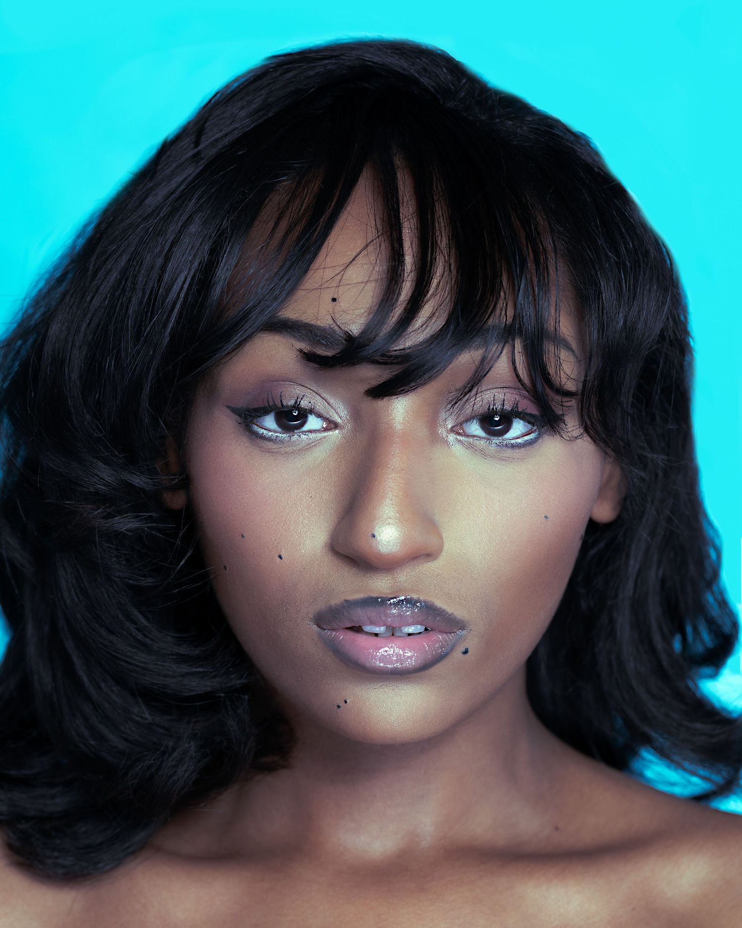 A young Black woman with black wavy hair and bangs poses against a cyan background with glittery lip makeup.