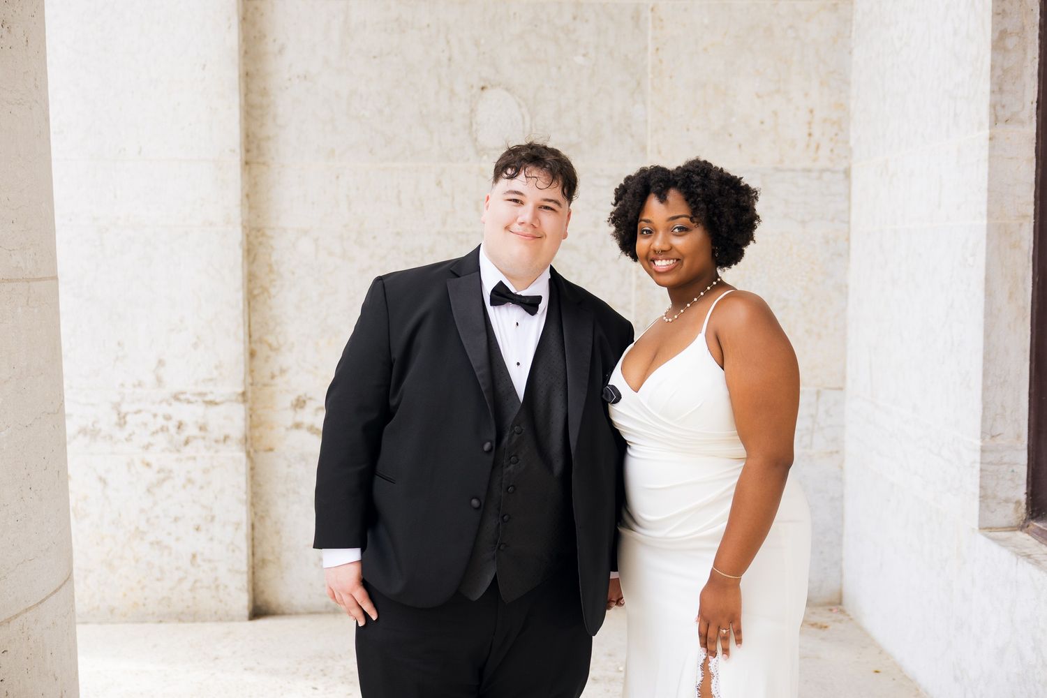 Couple in wedding attire, one in a black tuxedo and one in a white gown, smiling beside marble columns.