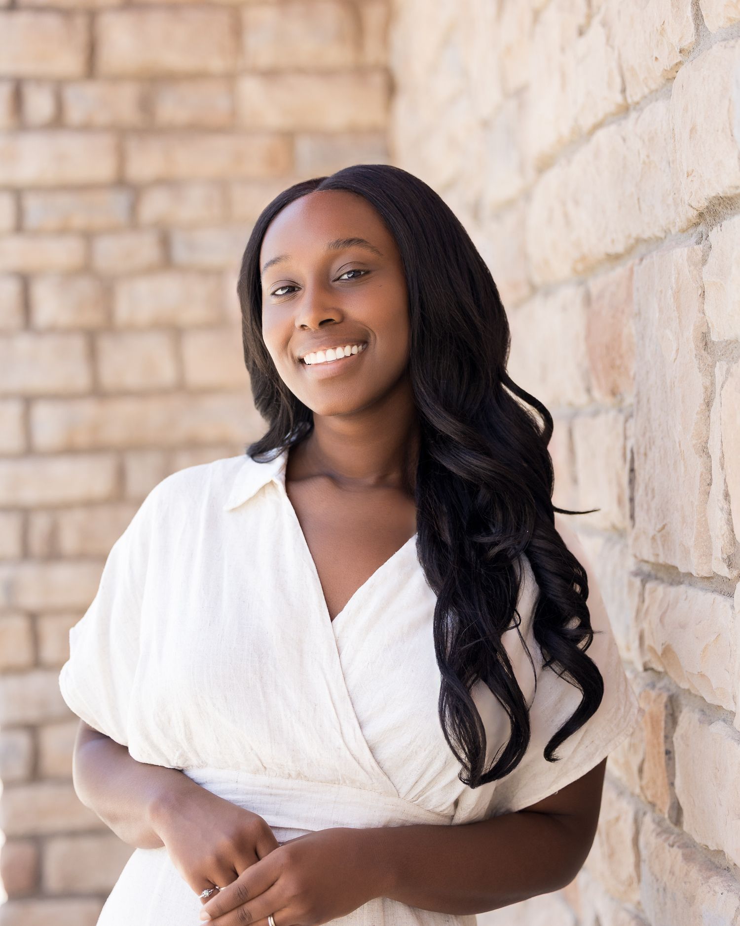 Smiling young woman with long black hair wearing a white wrap dress, leaning against a brick wall outdoors.