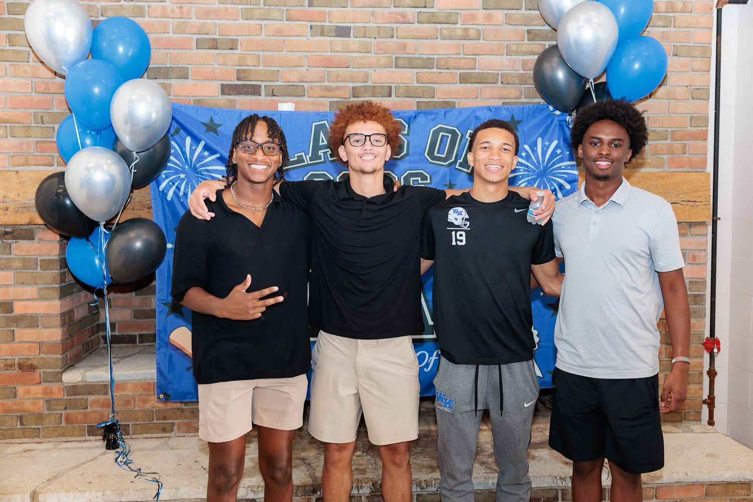 Four young men posing together in front of a blue banner with balloons at a school celebration event.