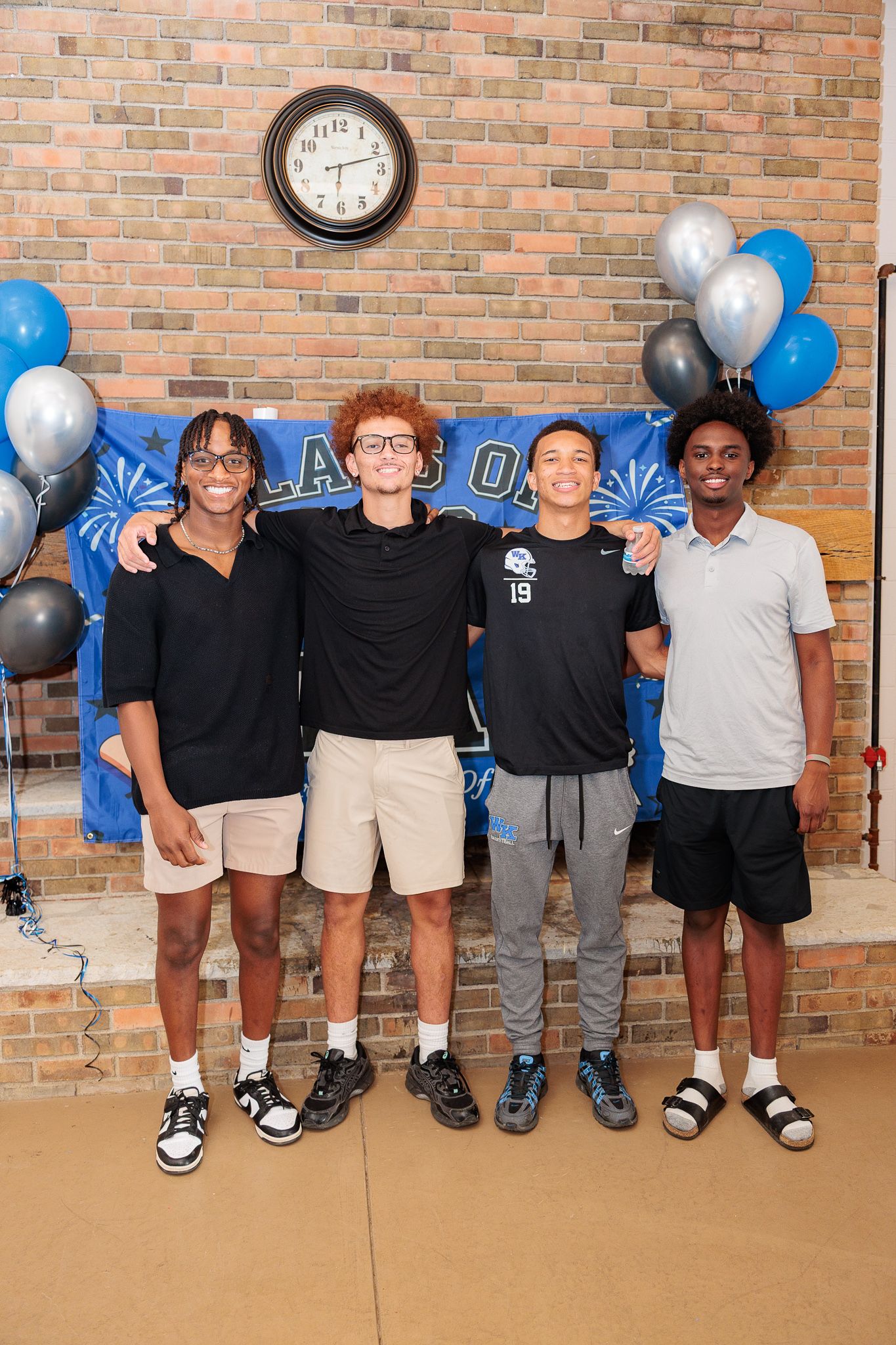 Four young men pose together smiling at a graduation celebration with blue and silver balloons and a Class of banner.