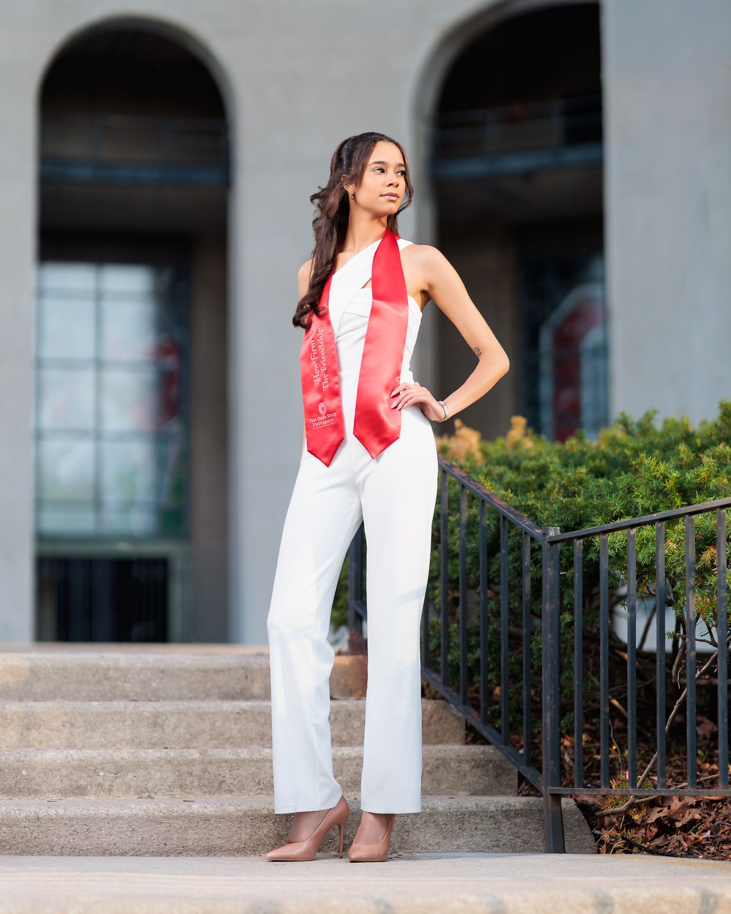 Young woman in white pantsuit and red graduation stole poses on outdoor steps near a building.