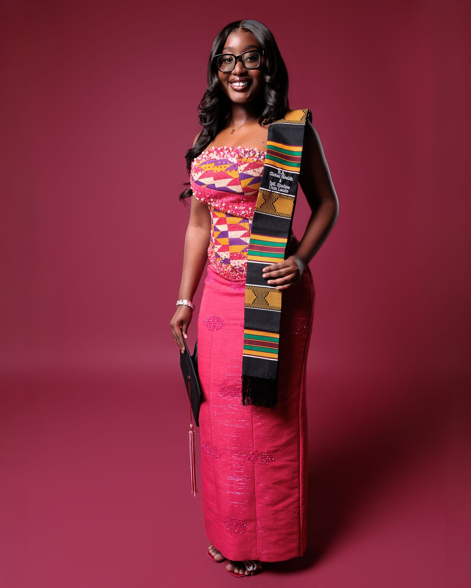 Woman in vibrant pink kente cloth dress and colorful sash posing against a deep red background.