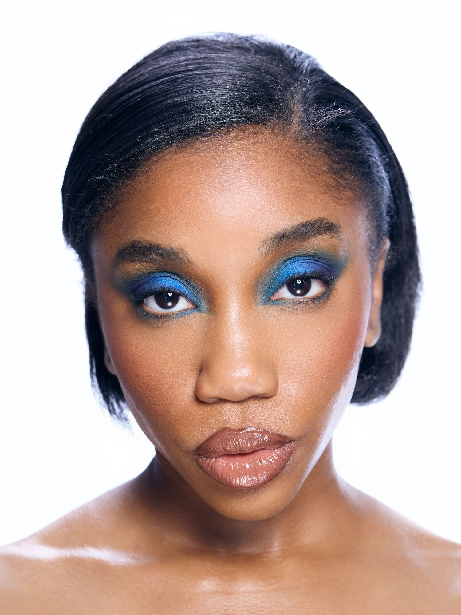 Woman with bold blue and green eyeshadow makeup, sleek black hair, and nude lips against white background.