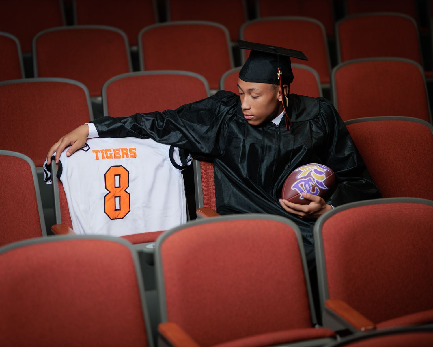 Graduate in cap and gown holds Tigers #8 jersey and football in auditorium seats during senior photo session.