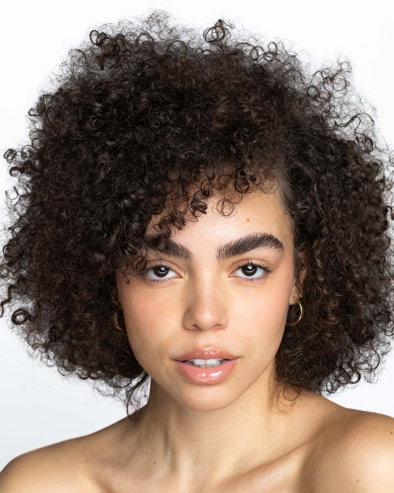 Young woman with voluminous dark curly hair, natural makeup, and gold hoop earrings against a white background.