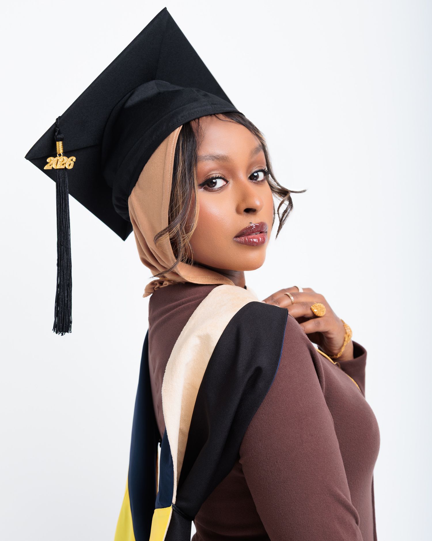 Young woman in graduation cap and gown posing confidently, wearing hijab, class of 2026 tassel, brown academic regalia.