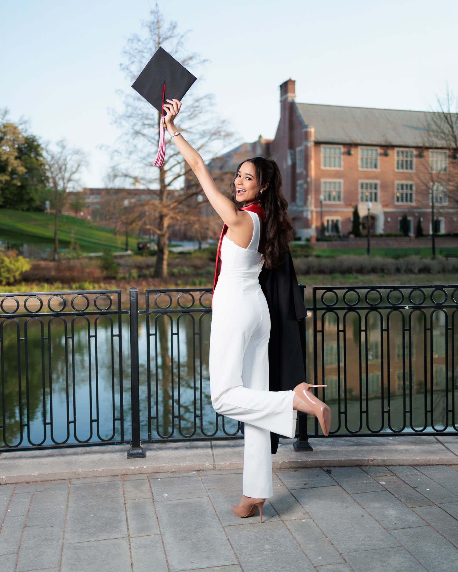 Graduating woman in white jumpsuit and black gown joyfully raising her cap by a campus pond.