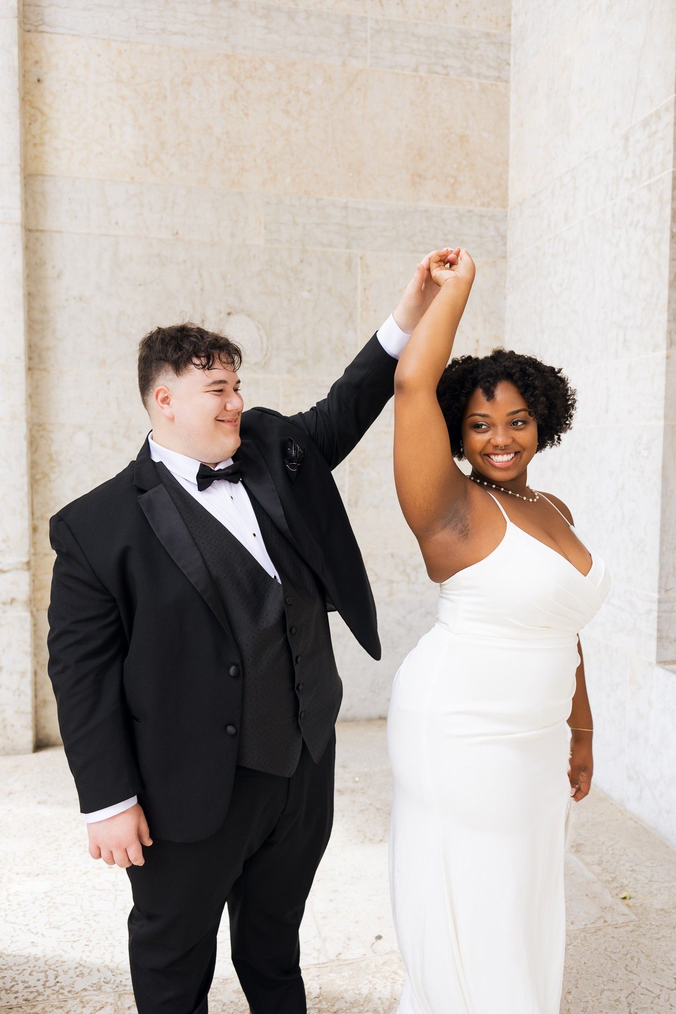 A cheerful wedding couple celebrates with raised hands, groom in black tuxedo spinning bride in elegant white dress outdoors.