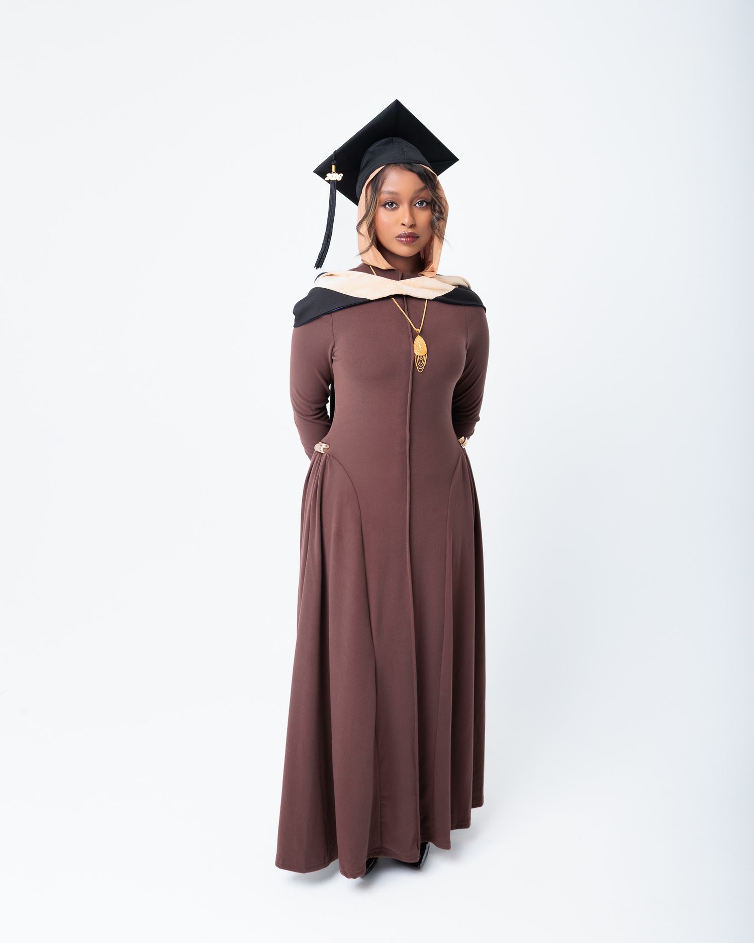 Young woman in brown graduation gown and black mortarboard cap poses proudly for formal graduation portrait.