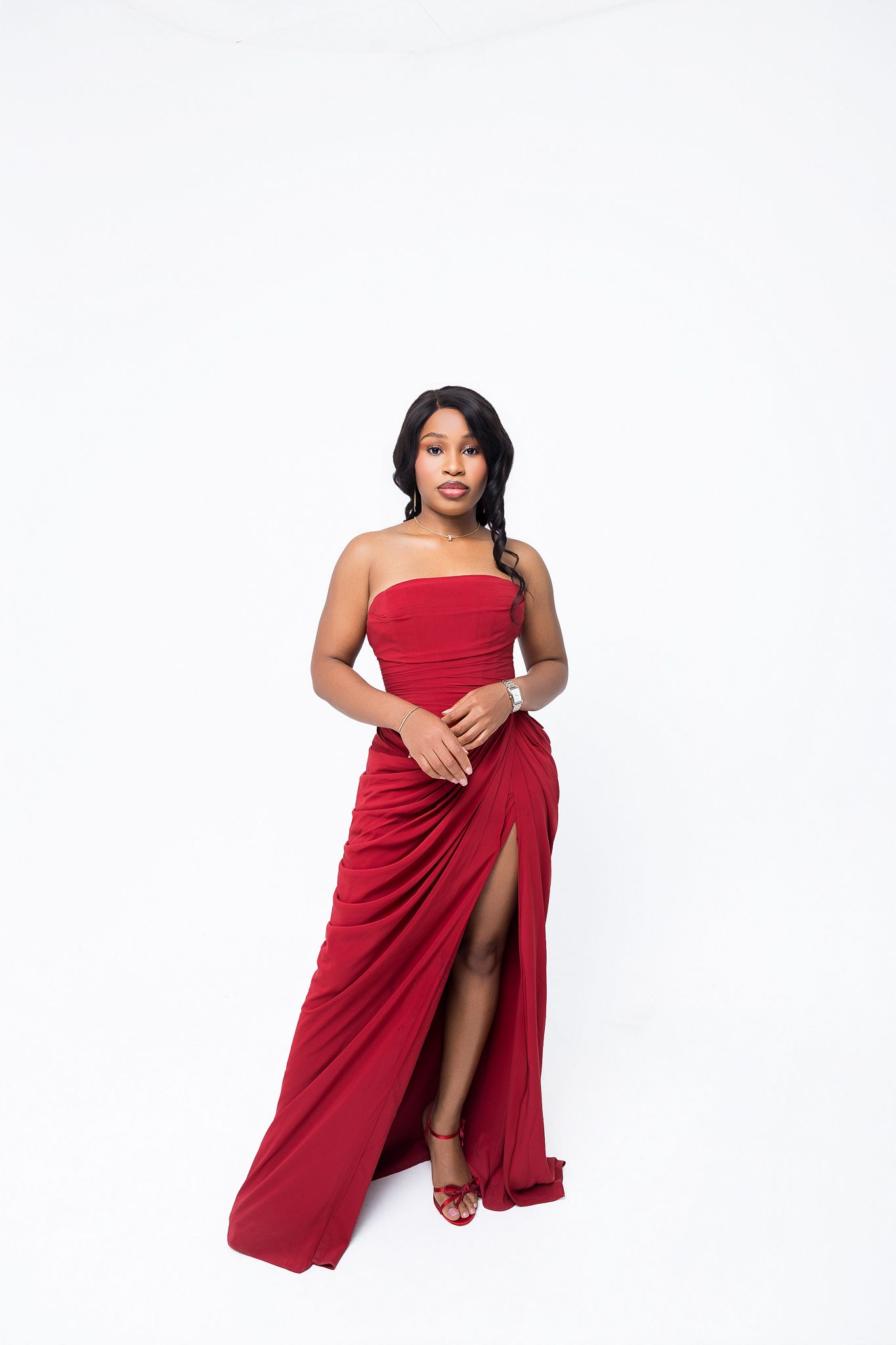 Woman in elegant red strapless gown with thigh-high slit and red heels posing against white background.