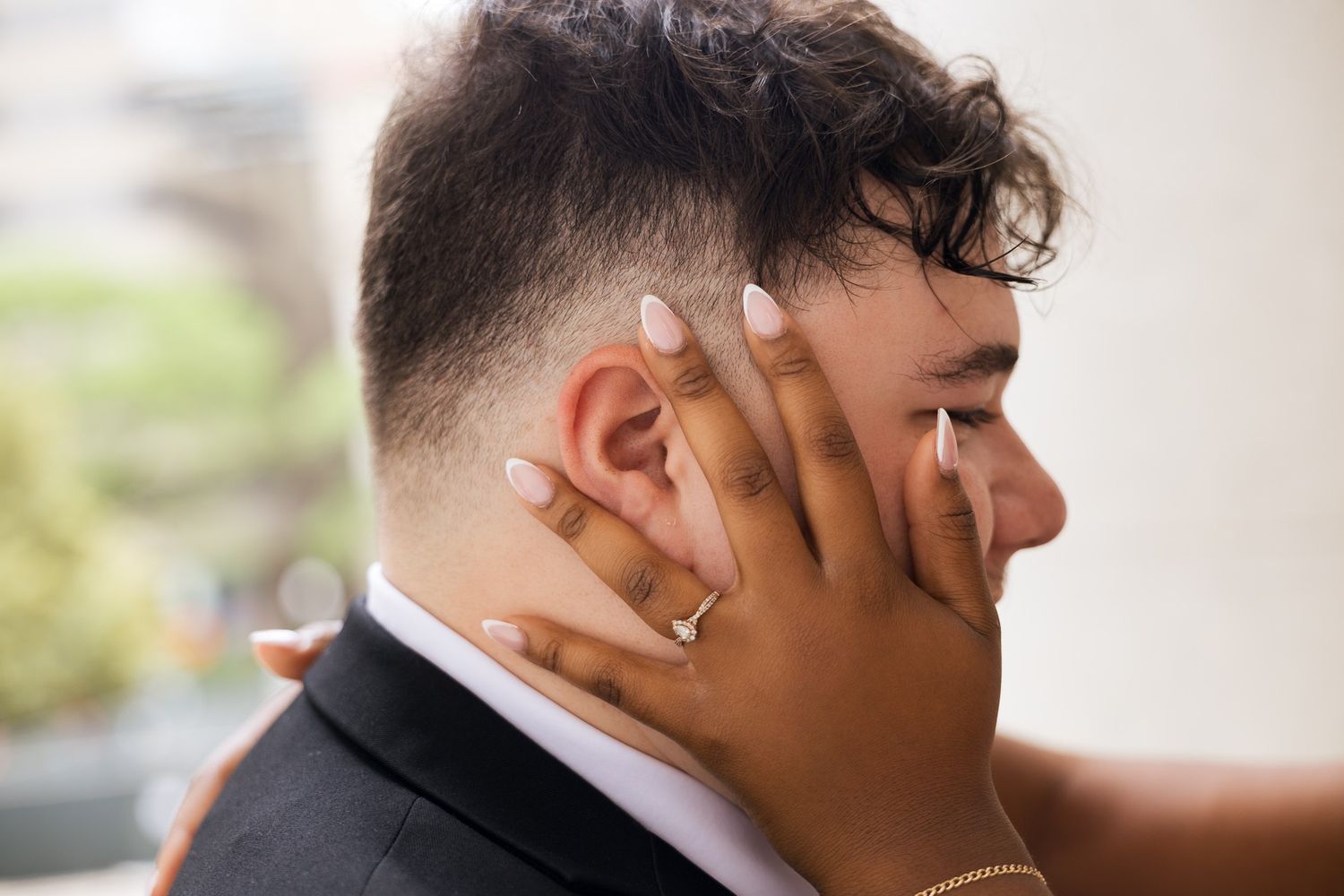A person with manicured nails and a gold ring gently cups the face of a man in a suit at a wedding.