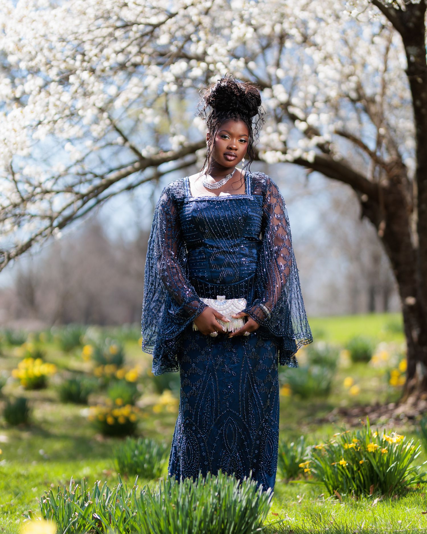 Young woman in navy beaded gown poses under blooming white tree surrounded by spring daffodils.