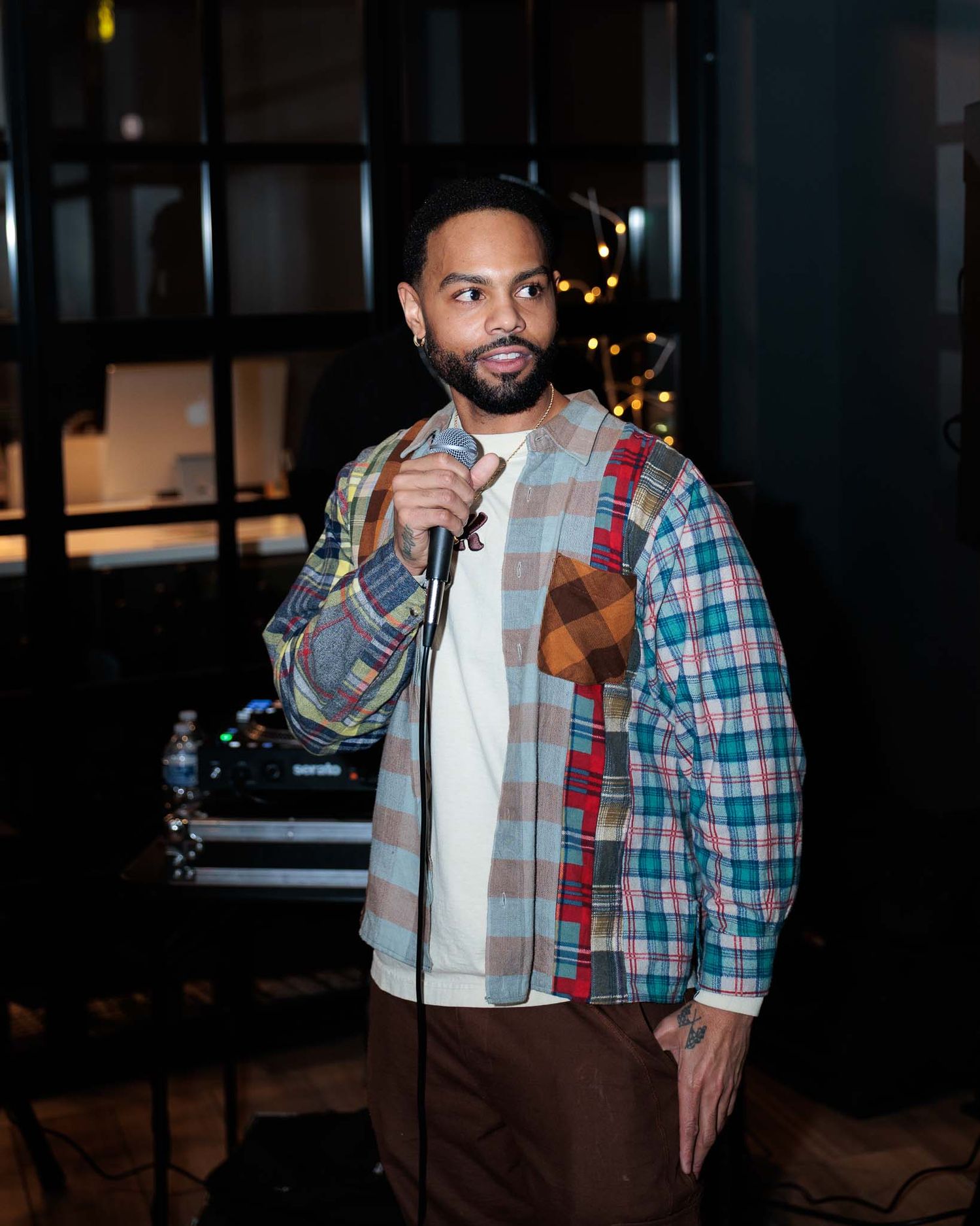 A person wearing a plaid shirt and light jacket stands in a dimly lit setting with blurred lights in the background.
