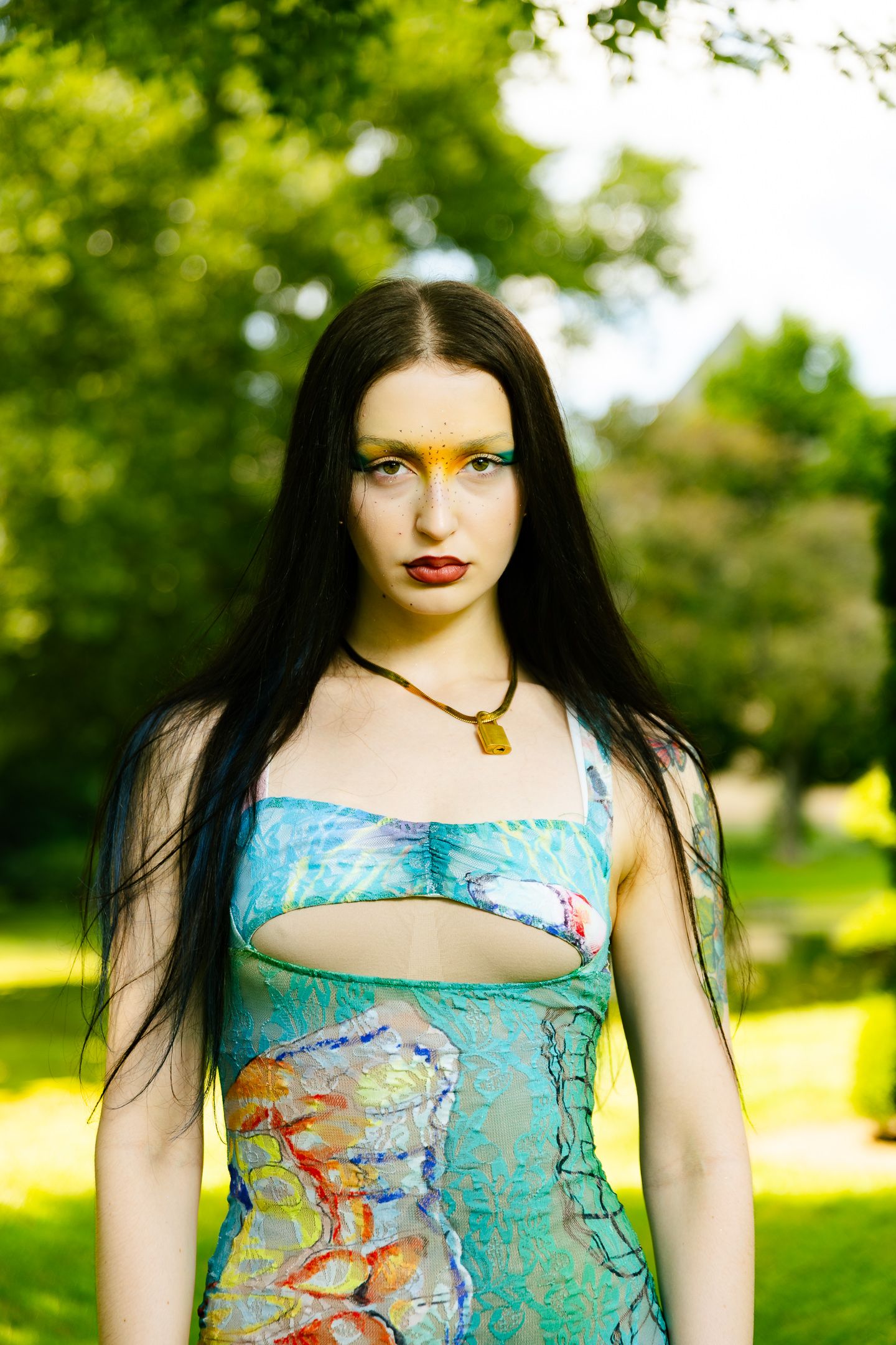 Woman in colorful aquatic print dress with artistic yellow eye makeup posing outdoors in a green park.