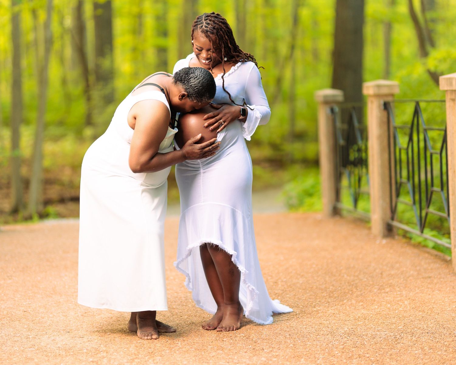 Two women in white dresses share a tender moment outdoors, one pregnant, embracing on a park path surrounded by green trees.