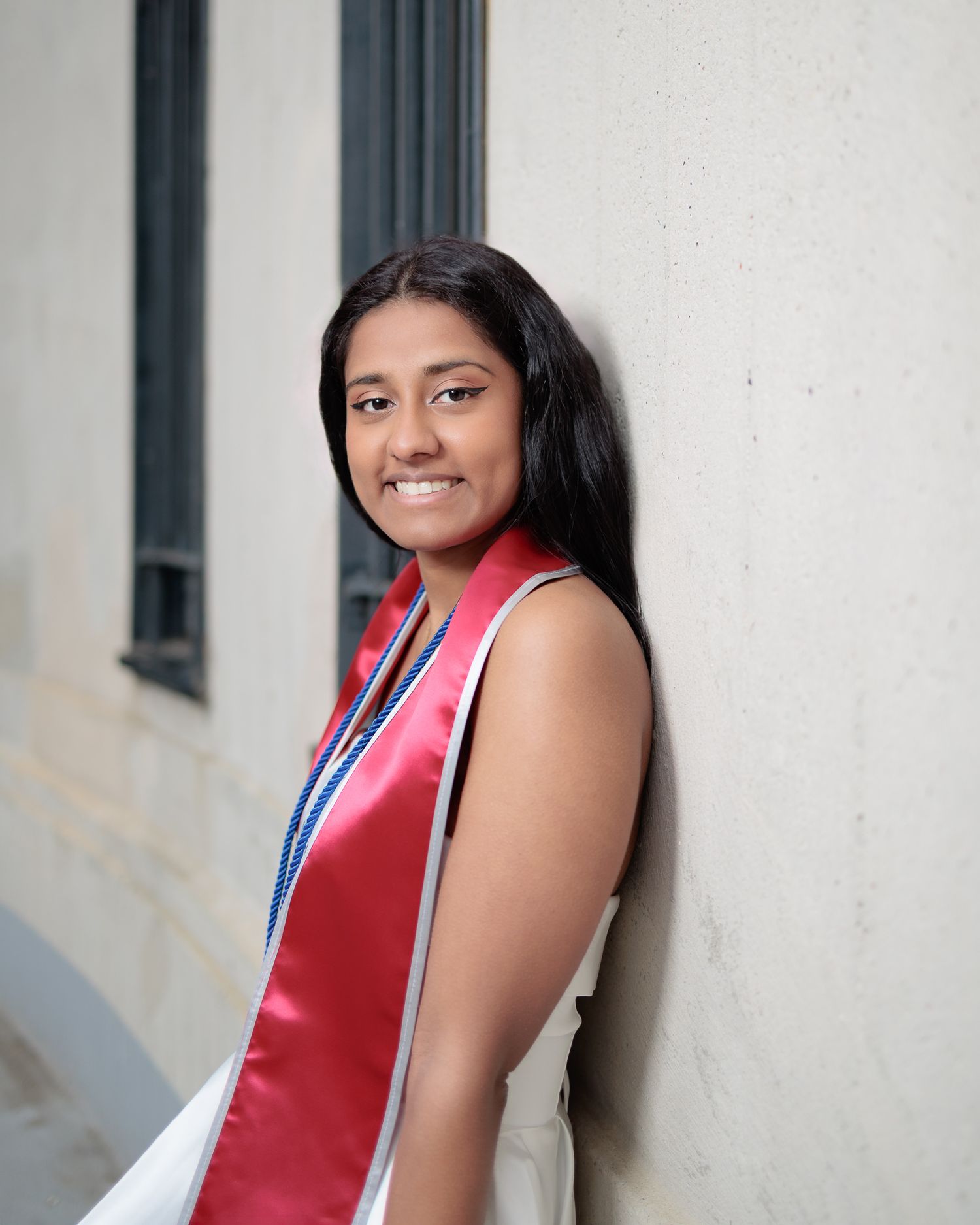 Smiling young woman in white dress with red graduation stole leaning against a wall outdoors.